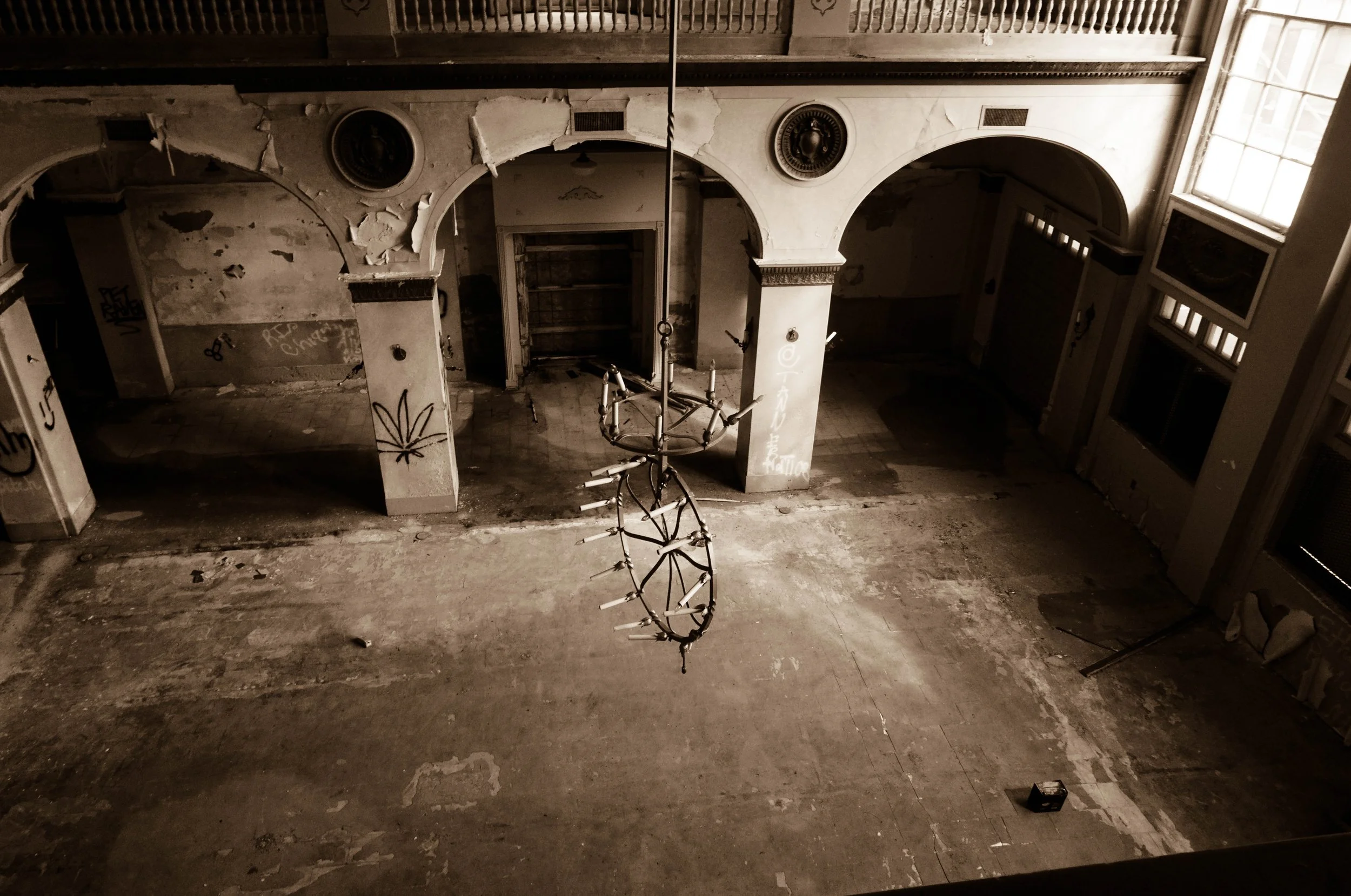 An abandoned, dilapidated room with peeling paint, graffiti, and broken windows, with a chandelier hanging from the ceiling.