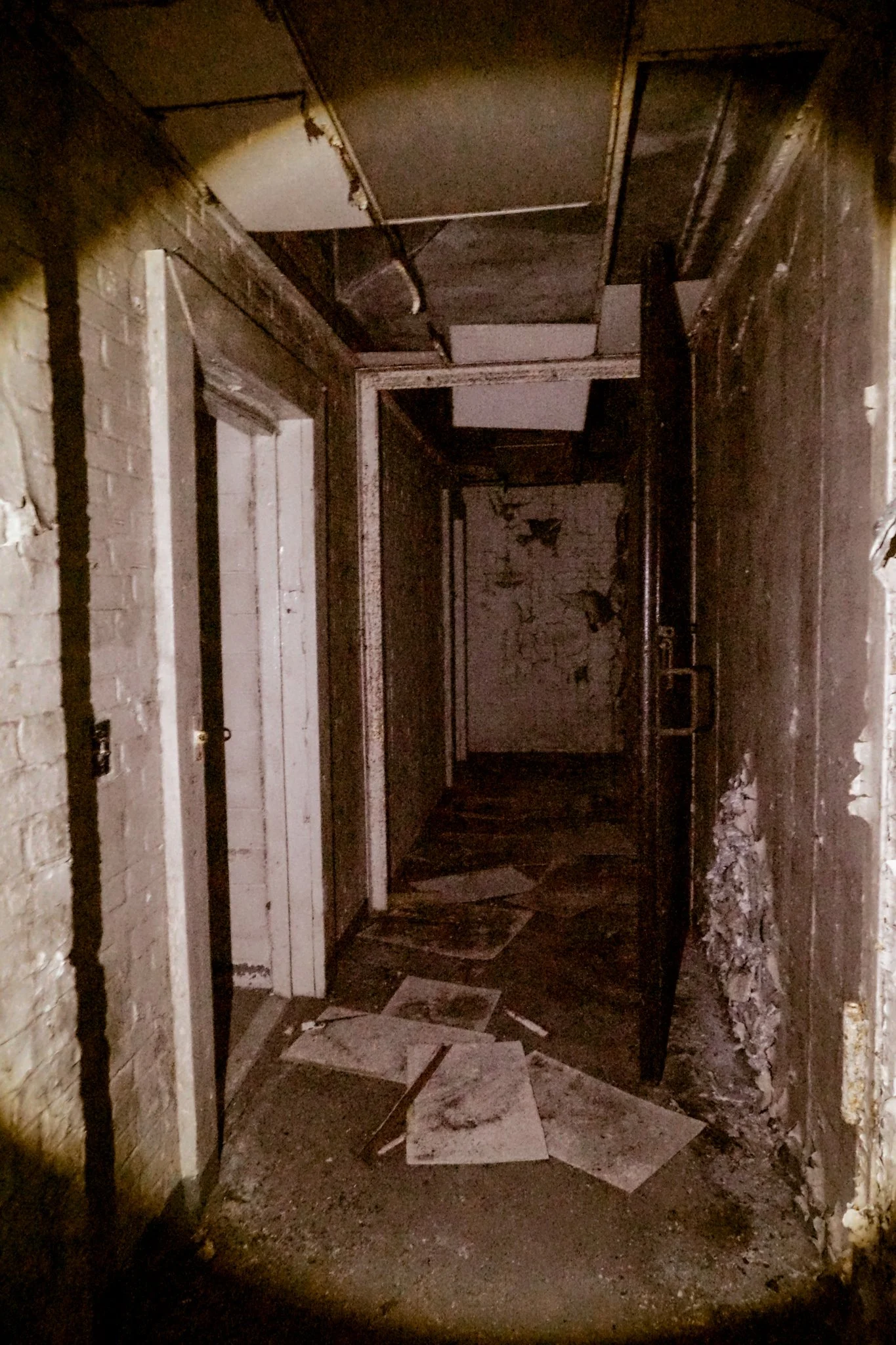 A dark, abandoned hallway with peeling paint, exposed pipes, and debris on the floor.