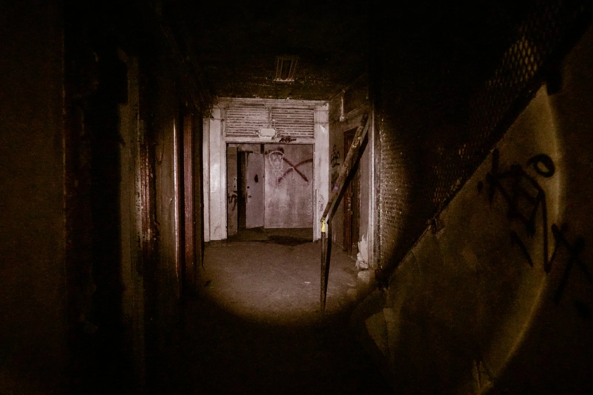 Dark, abandoned hallway with graffitied walls, a metal door at the end, and a broken trash can on the floor.