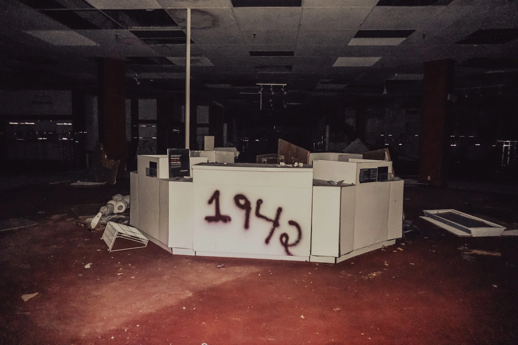 Abandoned electronics store with broken displays, scattered debris, graffiti reading '1942' spray-painted on a counter, dark interior and broken fixtures.