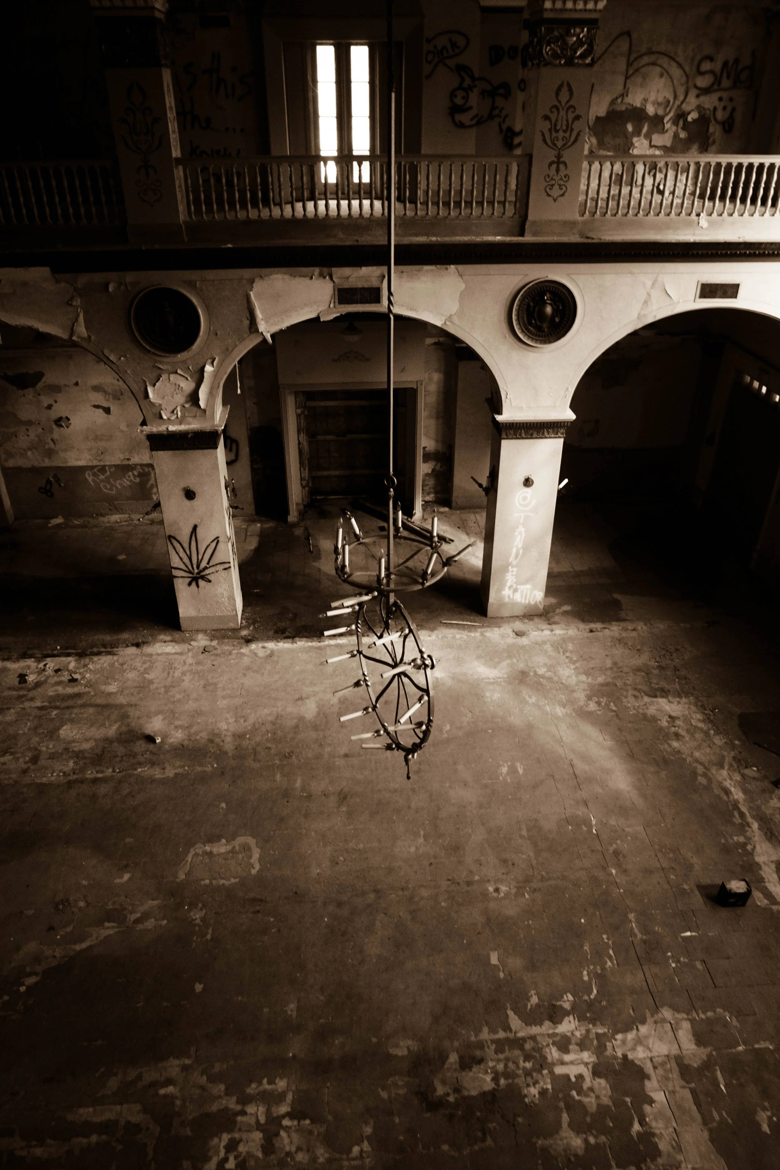 Abandoned, dilapidated room with peeling paint, graffiti, and broken fixtures. A chandelier hangs upside down from the ceiling.