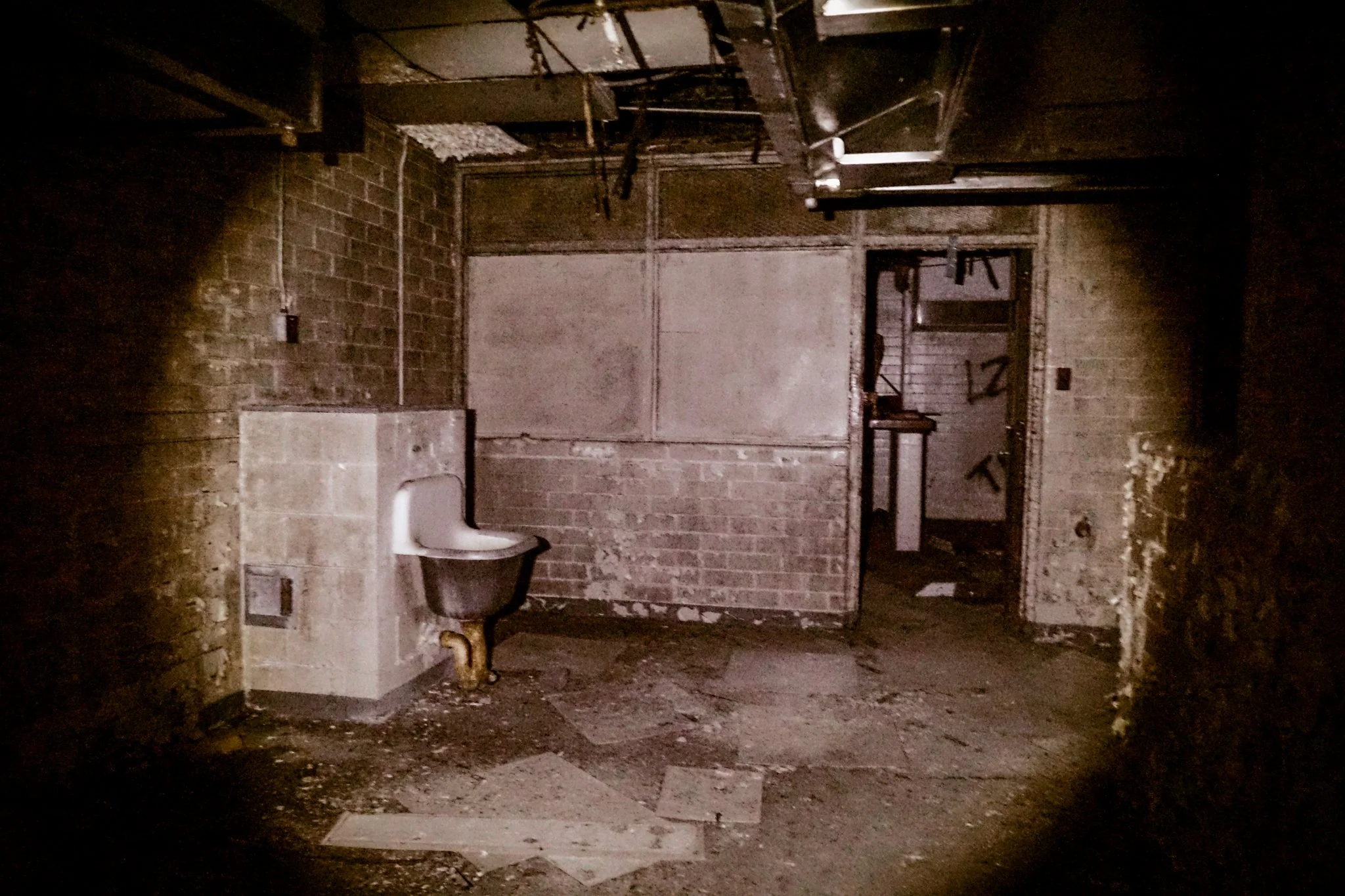 An abandoned room with exposed brick walls, a broken floor, and graffiti on the doorway. There is a dirty toilet against the wall, and ceiling pipes and wires hanging down.