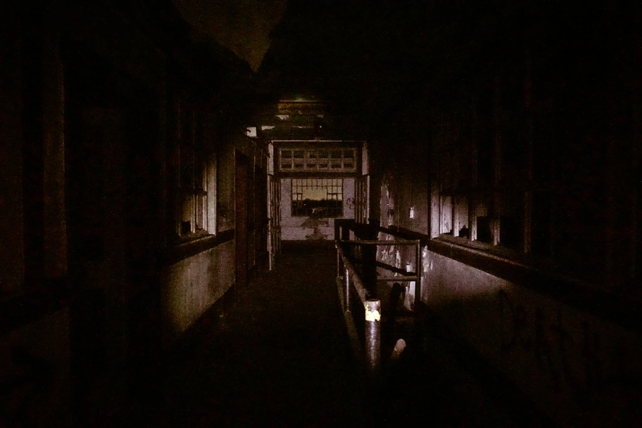 Dark, abandoned hallway with broken railings and windows, faint light coming from the doorway at the end, possibly from outside, creating minimal illumination in the eerie scene.