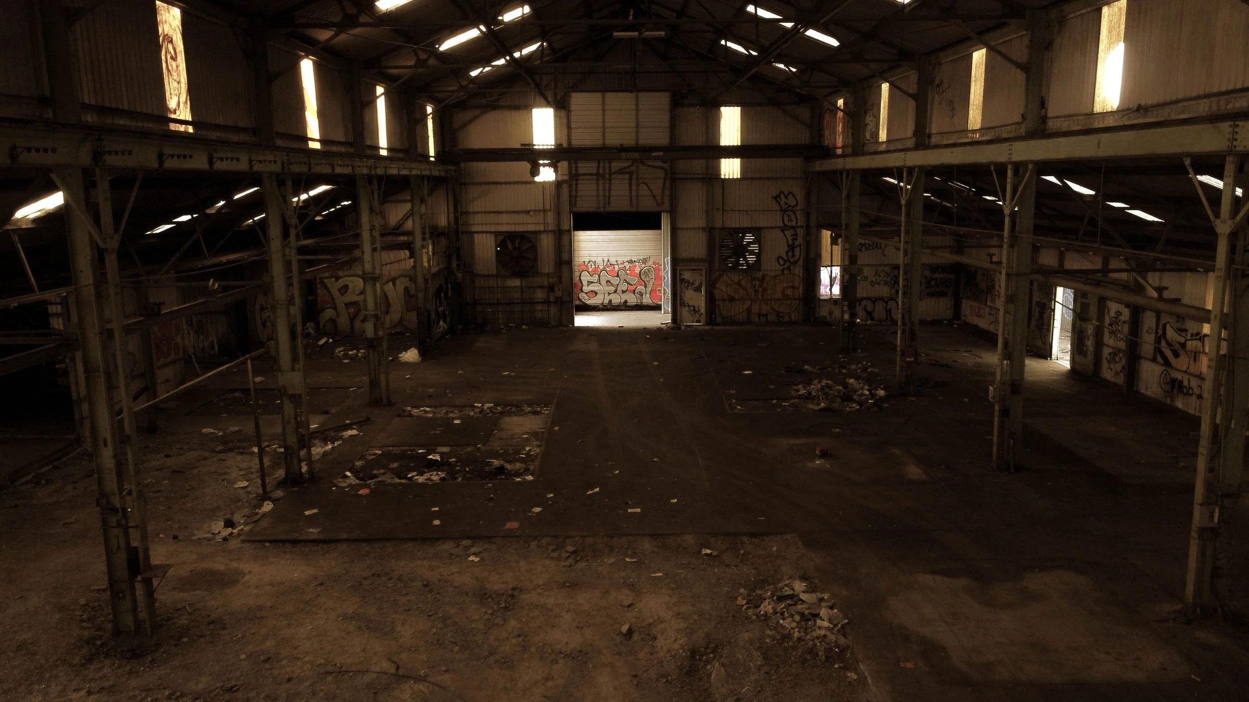 Interior of an abandoned warehouse with graffiti on the walls, dirt-covered floor, and some debris, illuminated by limited natural light from small windows and an open garage door.