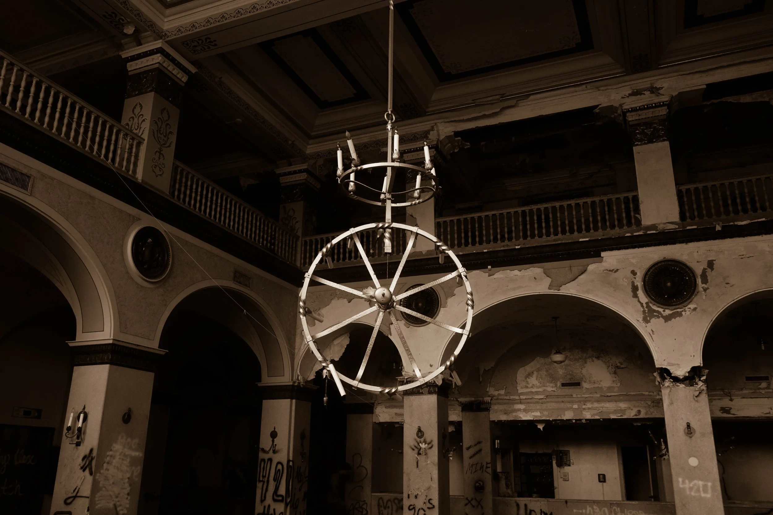 An abandoned, dilapidated building with peeling paint, graffiti on the columns, and broken chandeliers hanging from the ceiling.