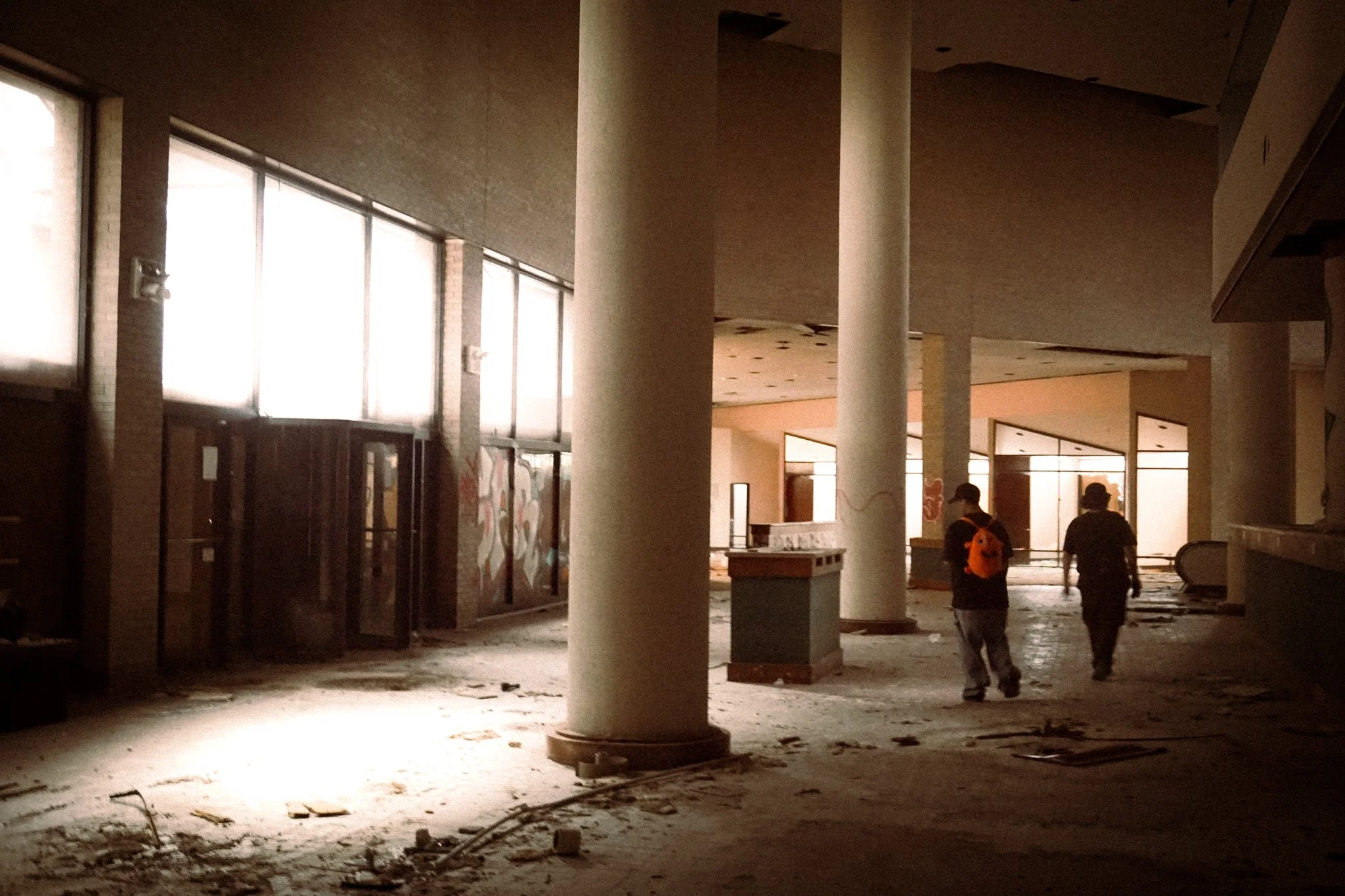The image shows an abandoned, dimly lit indoor space with large windows, concrete columns, and debris on the floor. Two people are walking through the area, carrying backpacks and dressed casually.