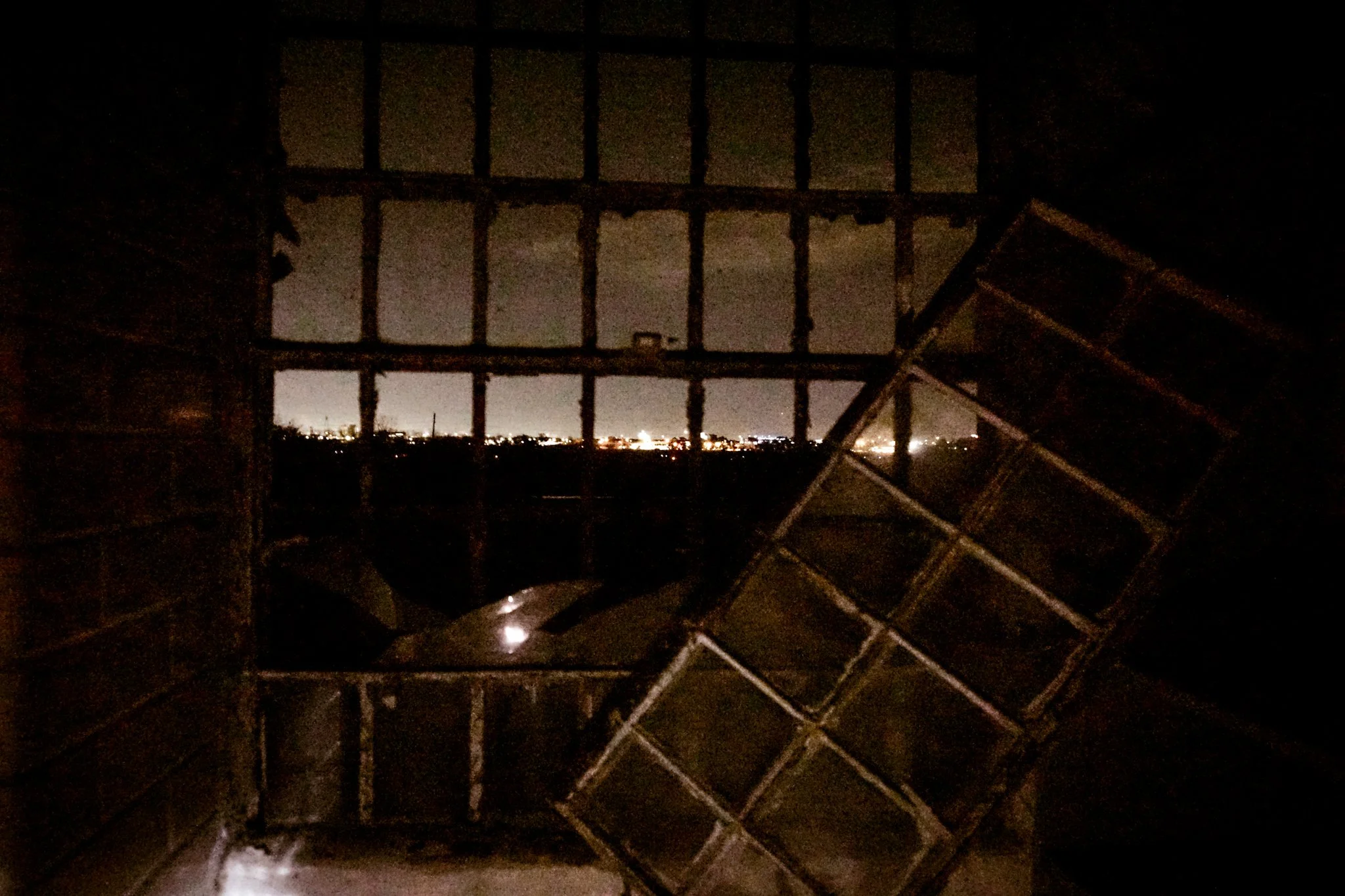 Nighttime cityscape seen through an open, boarded-up window with an angled kitchen rack in the foreground.