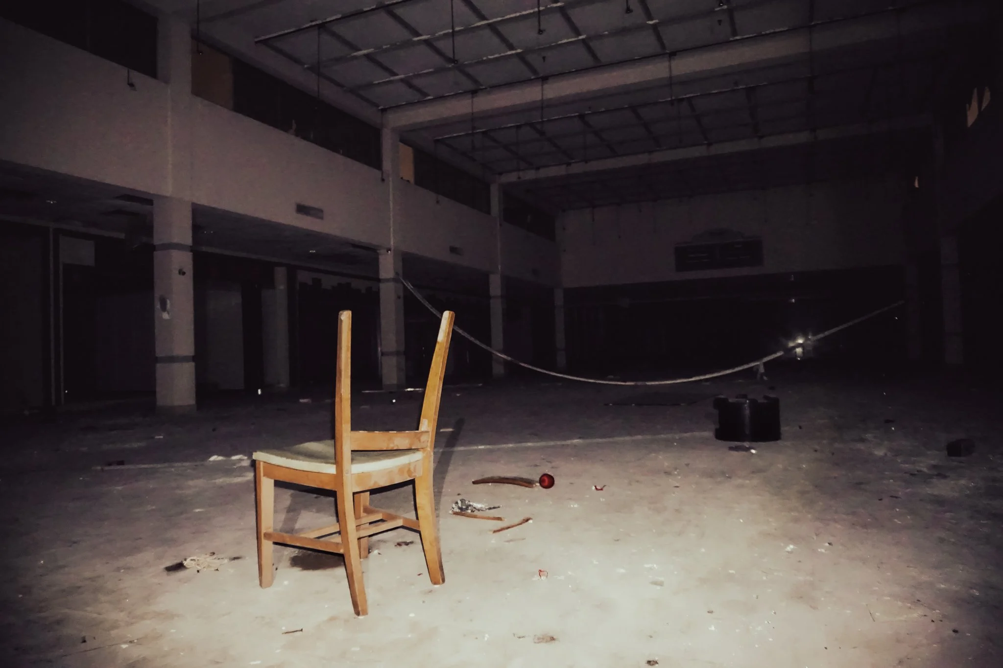 An abandoned, dark building interior with an empty wooden chair in the foreground. The space is dimly lit, with debris scattered across the dusty floor, and a damaged rope or wire hanging loosely.