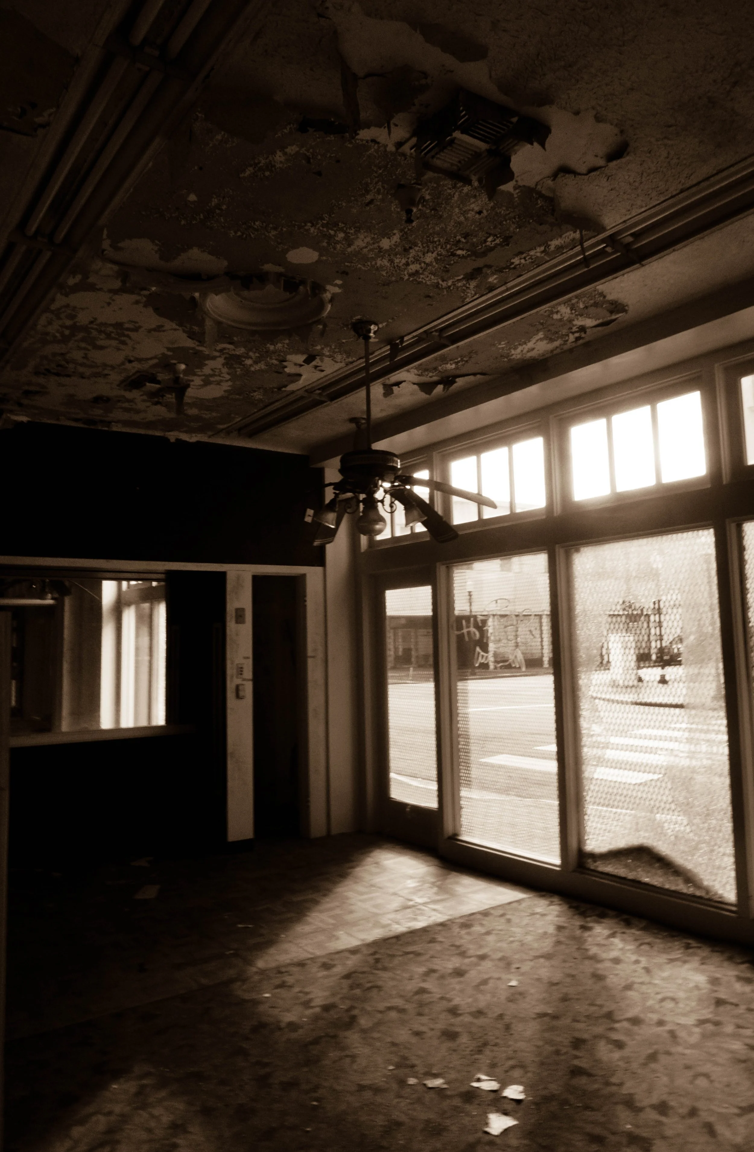 A dimly lit, abandoned room with a peeling ceiling, a ceiling fan, and large windows with sunlight streaming in.