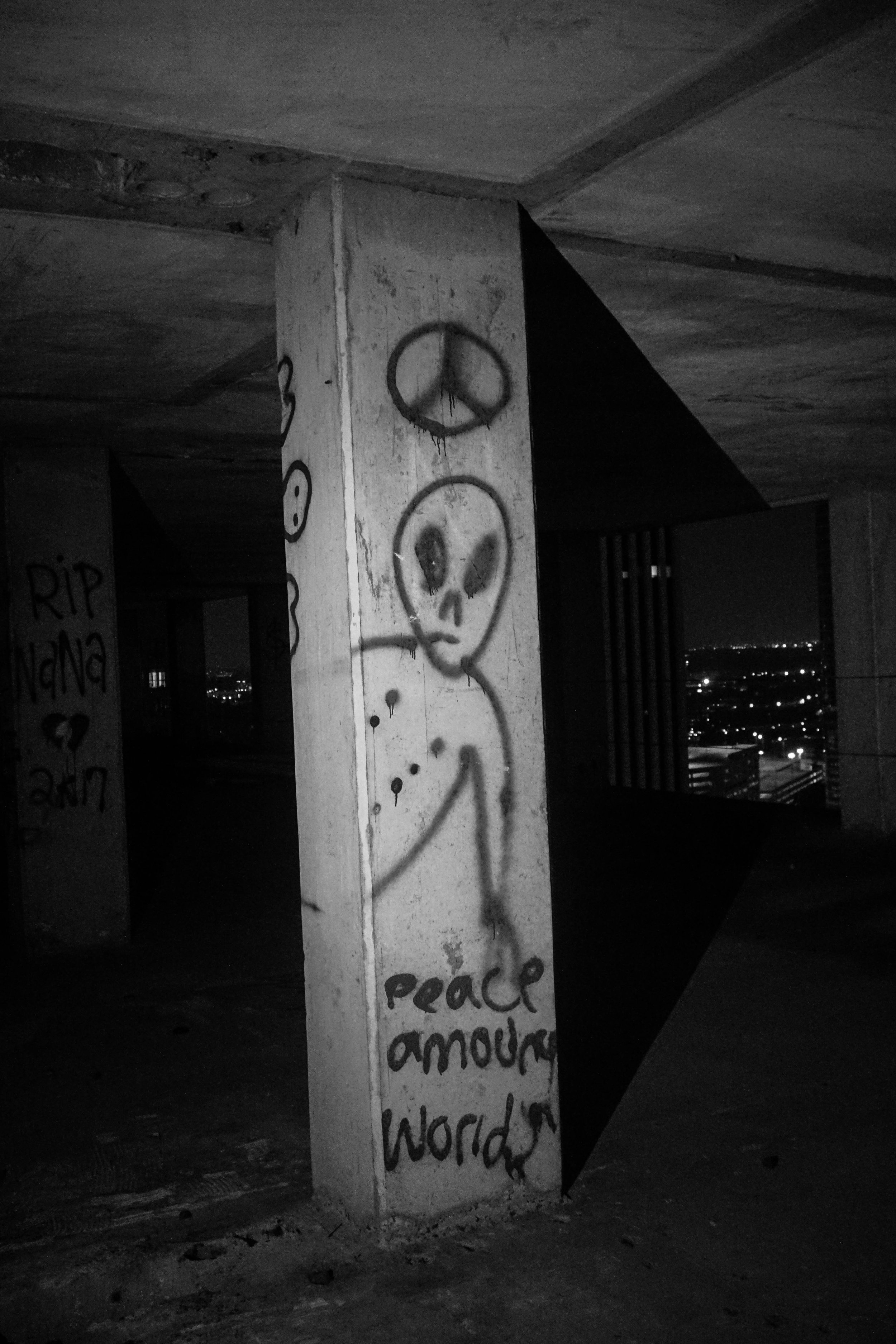 Graffiti of an alien figure with a peace sign above its head and the words "peace amour worlds" written below on a concrete pillar in a dark, empty underground parking garage at night.