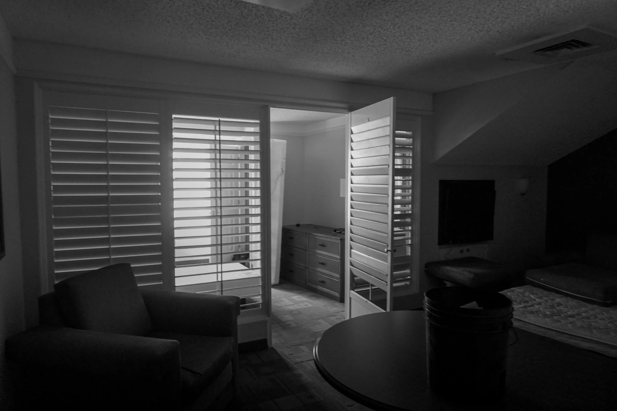 Dimly lit room with an open door leading to a balcony or patio area, featuring shutters on the window and door. Inside the room, there is a chair, a round table with a bucket, and a bed with bedding and pillows. A dresser and a wall-mounted TV are vi