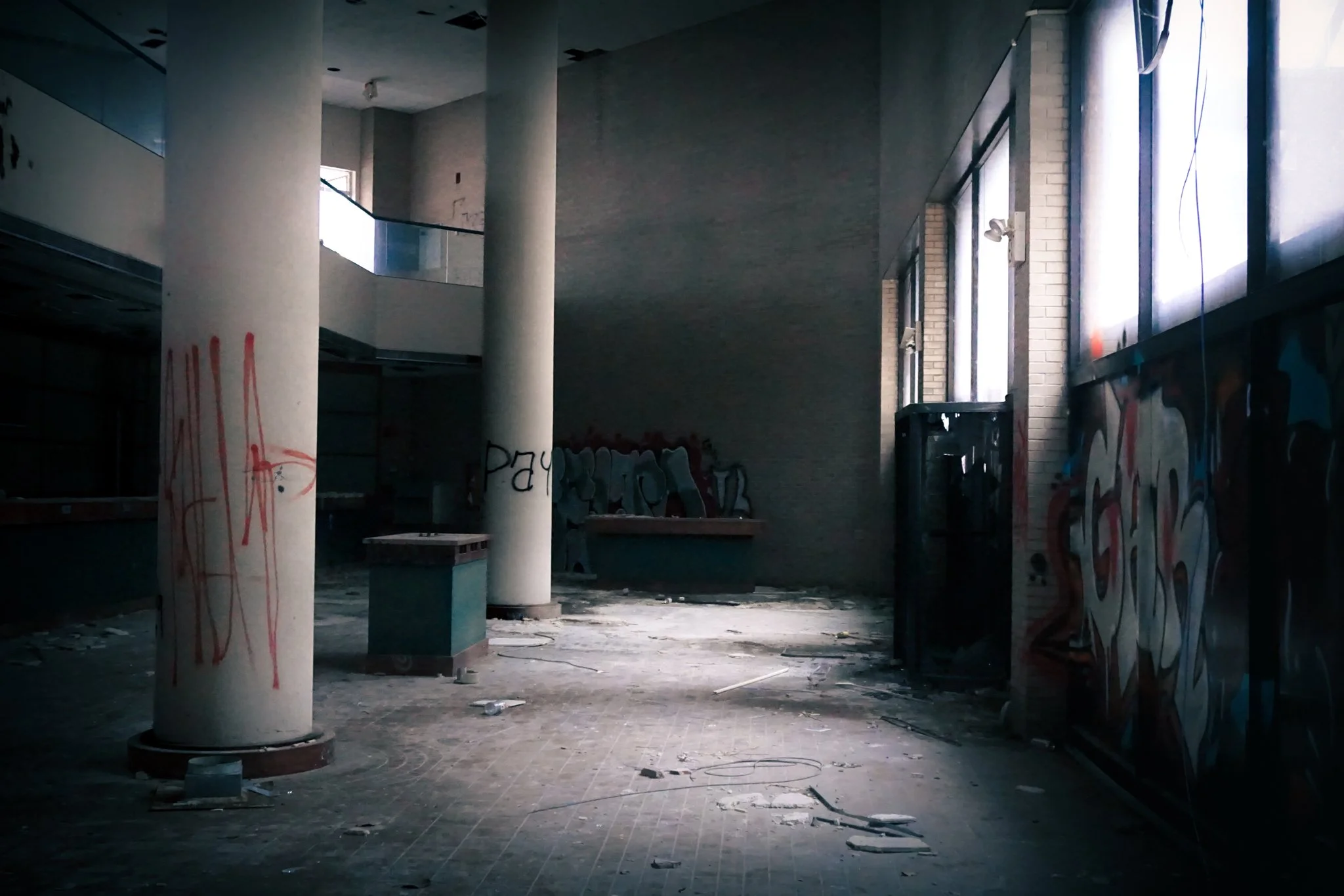Interior of an abandoned building with graffiti on columns and walls, debris on the floor, and large windows allowing natural light to enter.