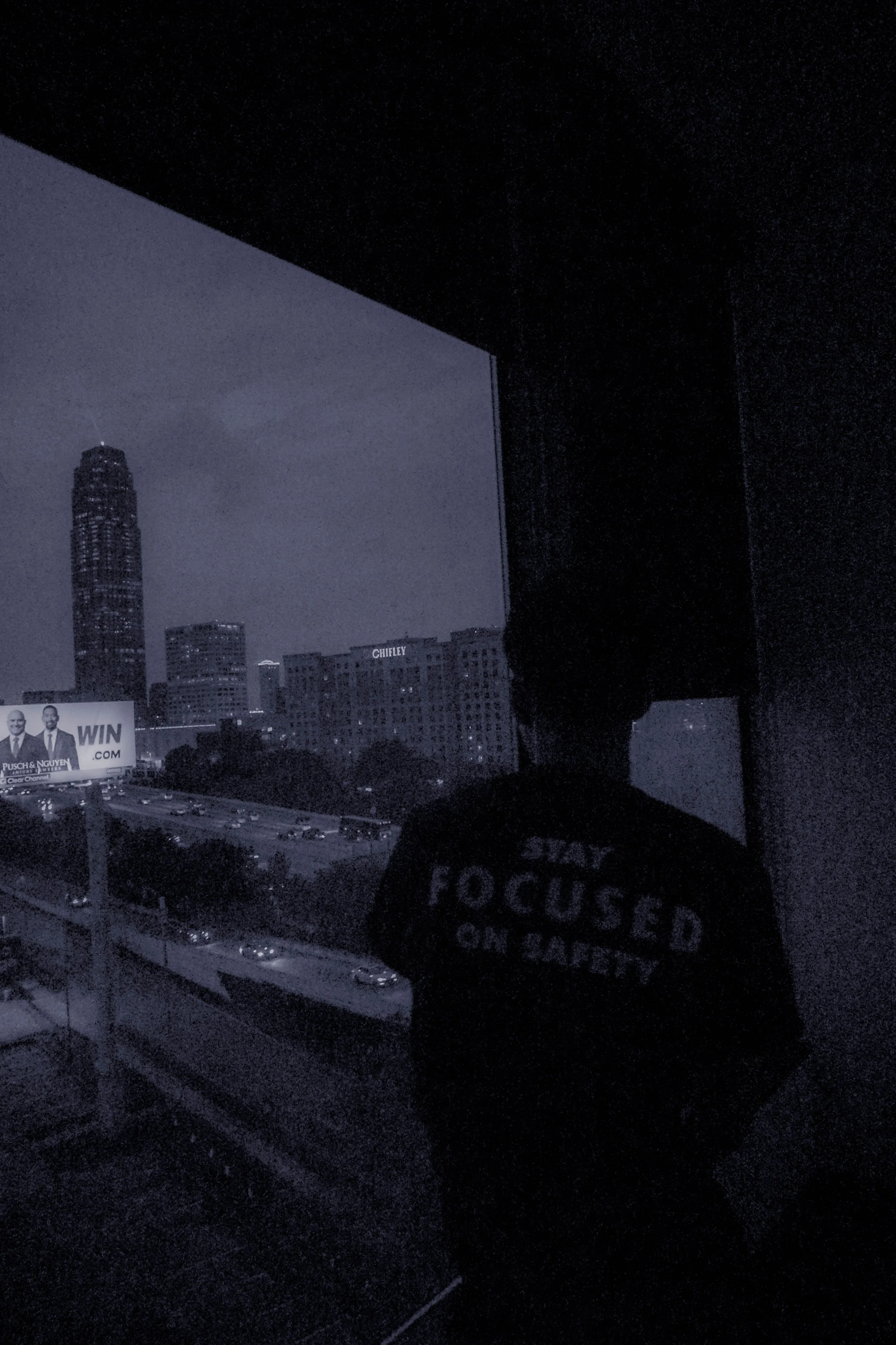 A person standing inside a dark room facing a large window showing a cityscape at night, with tall buildings and a billboard in the distance. The person is wearing a dark T-shirt with white text that reads 'FOCUSED ON CHOICE'.