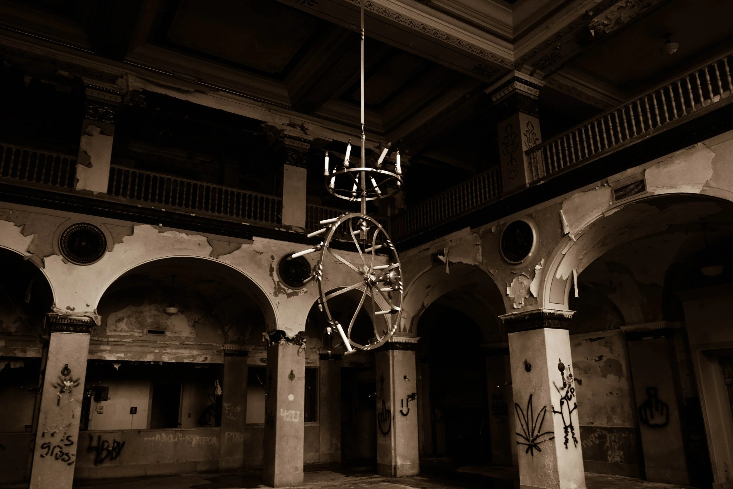 An abandoned, dilapidated interior with peeling paint, graffiti on walls, and broken chandeliers hanging from the ceiling.