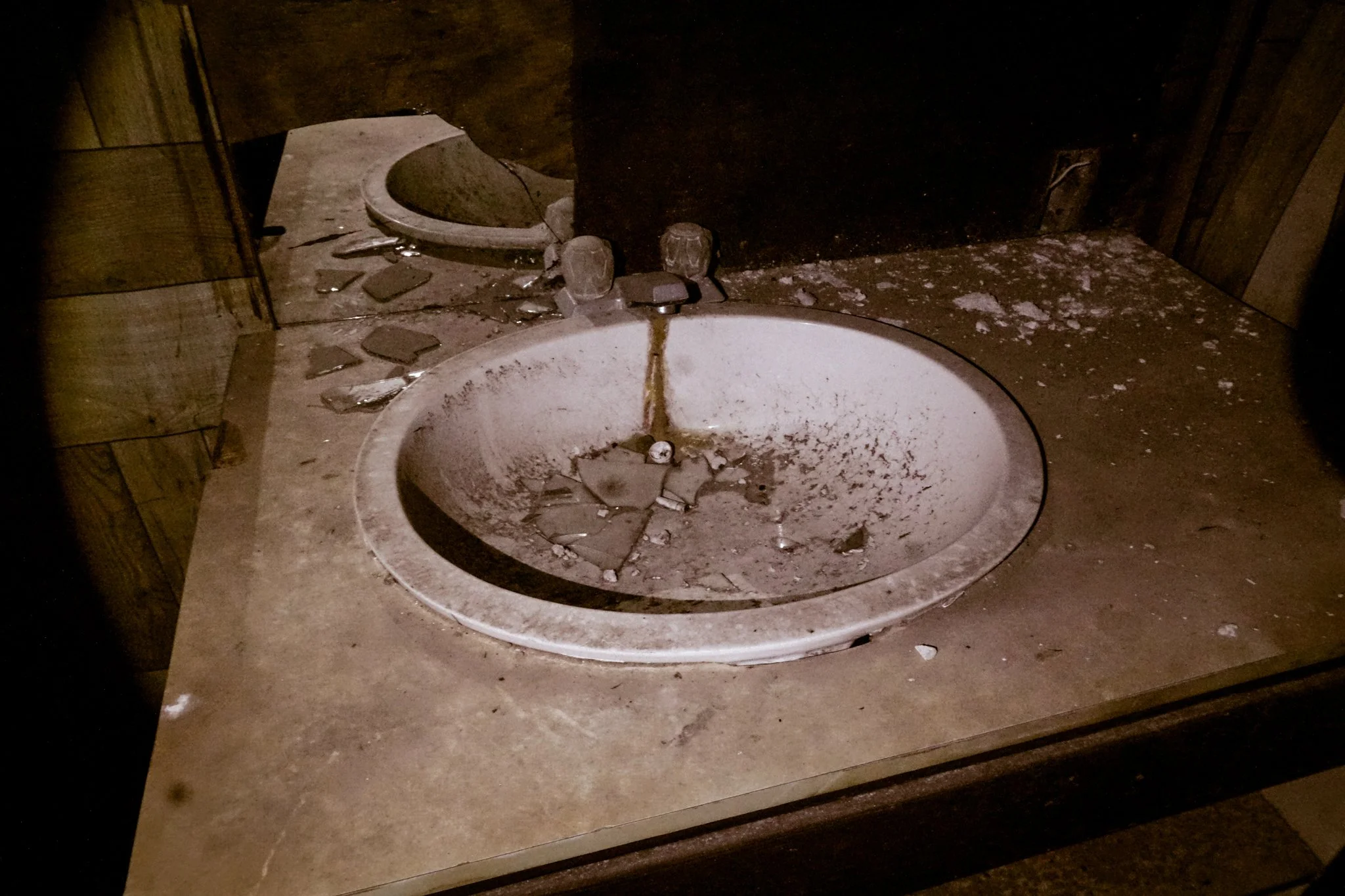 Dirty and broken bathroom sink with debris and shattered tiles on the countertop, and a cracked mirror in the background.