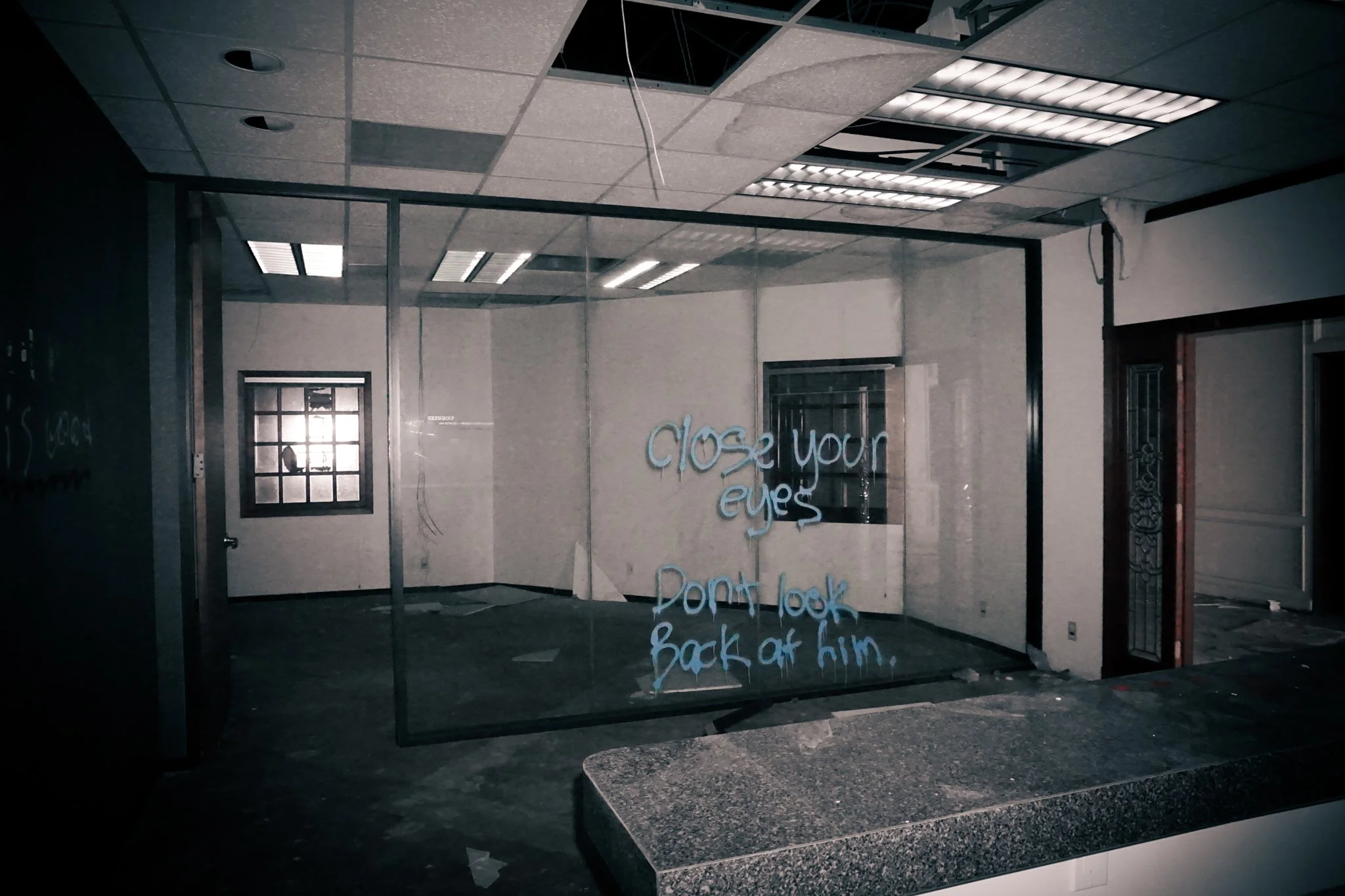 Empty office space with dirty floors, broken ceiling tiles, and a glass wall with a handwritten message in blue ink that says, 'close your eyes don't look back at him.'