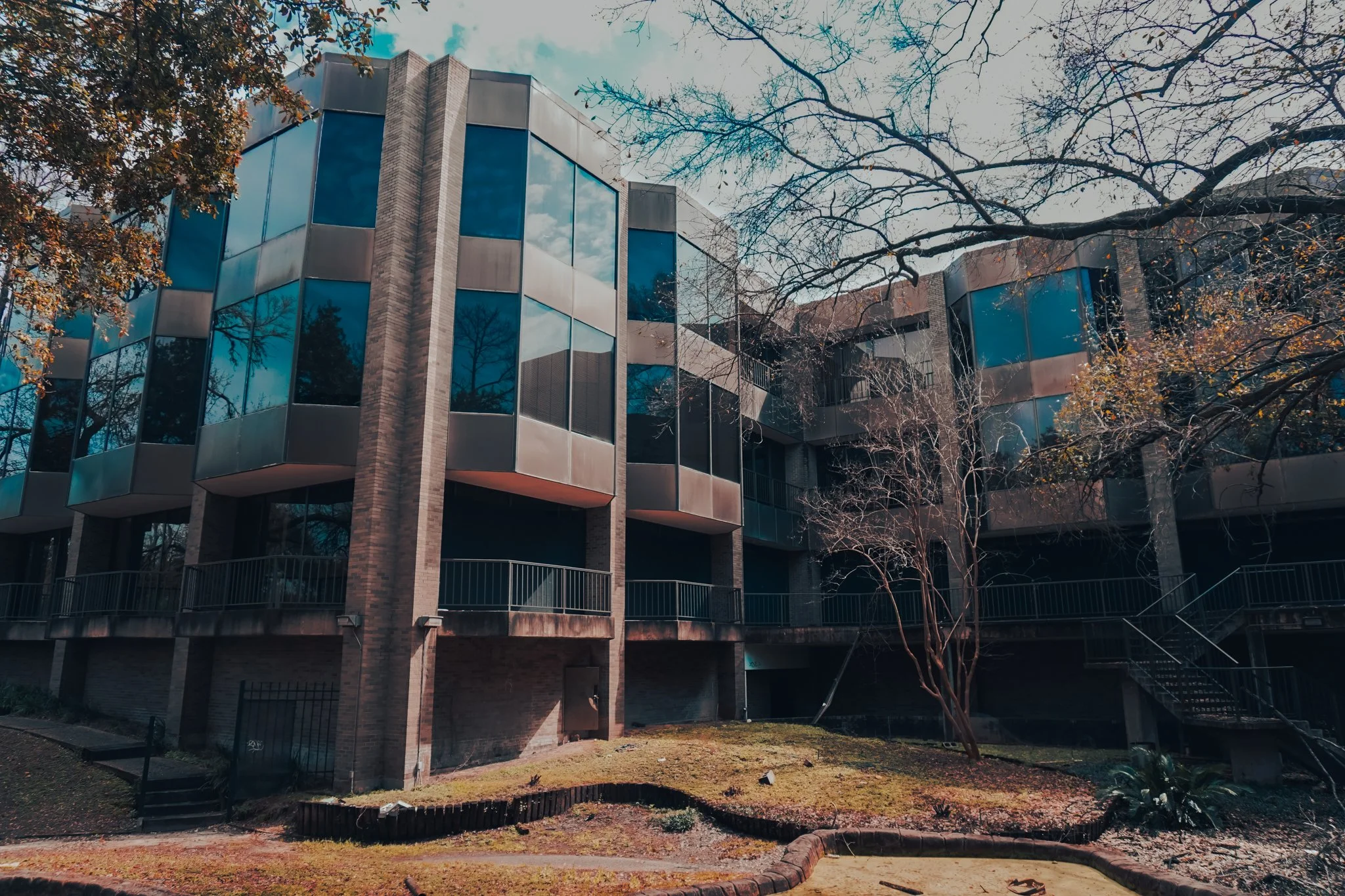 A modern multi-story building with large reflective blue glass windows and brick walls. The building is surrounded by trees, some with bare branches, and the scene appears to be during late fall or winter. There is a grassy area in the foreground wit