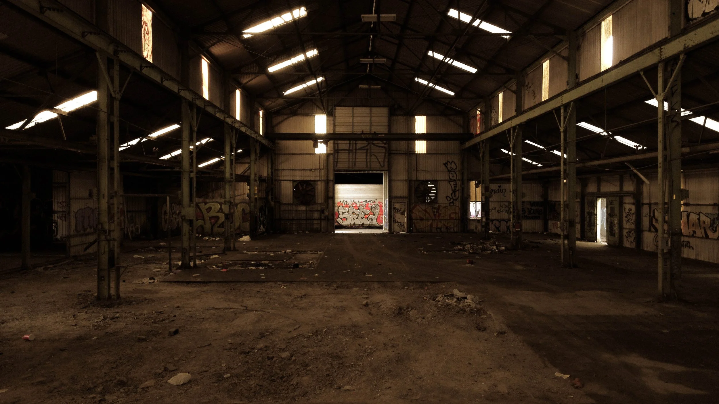An empty, abandoned warehouse with metal structure and graffiti on the walls, dirty floor with scattered trash, and sunlight streaming through the broken panels of the roof.