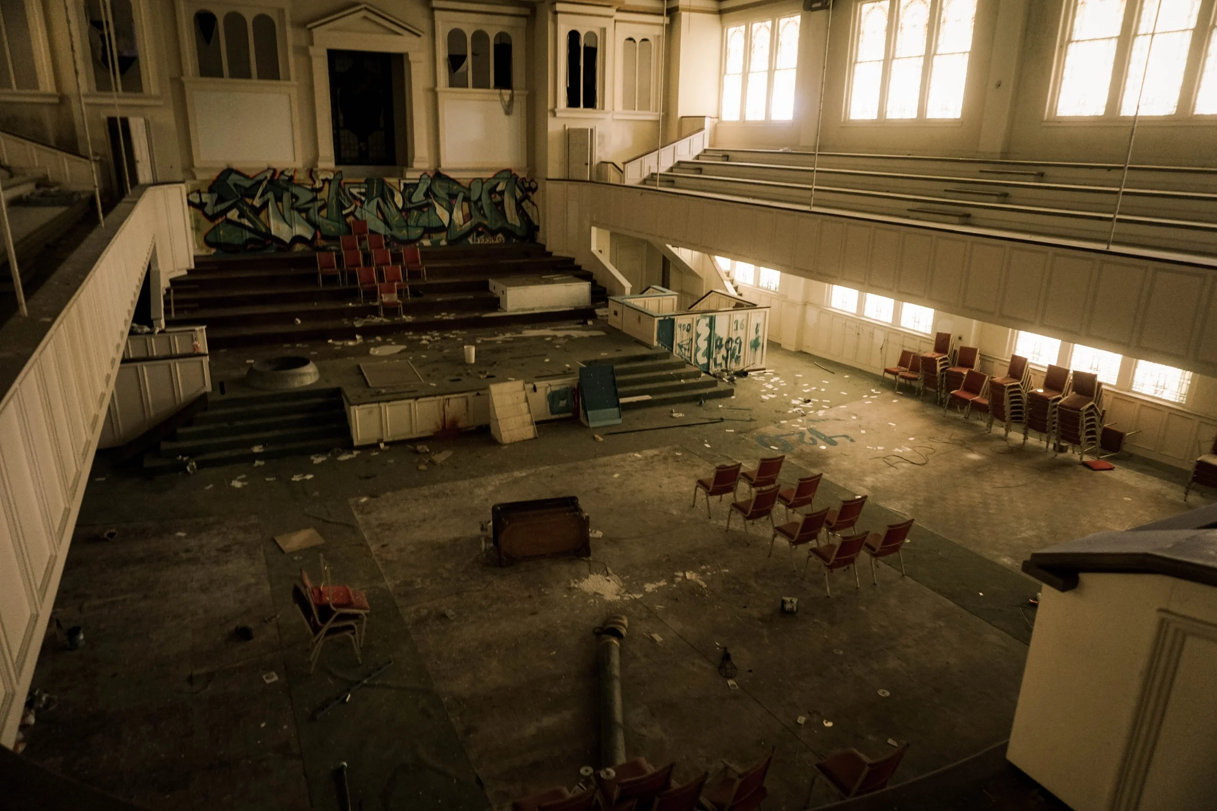 An abandoned, ruined auditorium with scattered chairs, debris on the floor, and graffiti on the stage area.