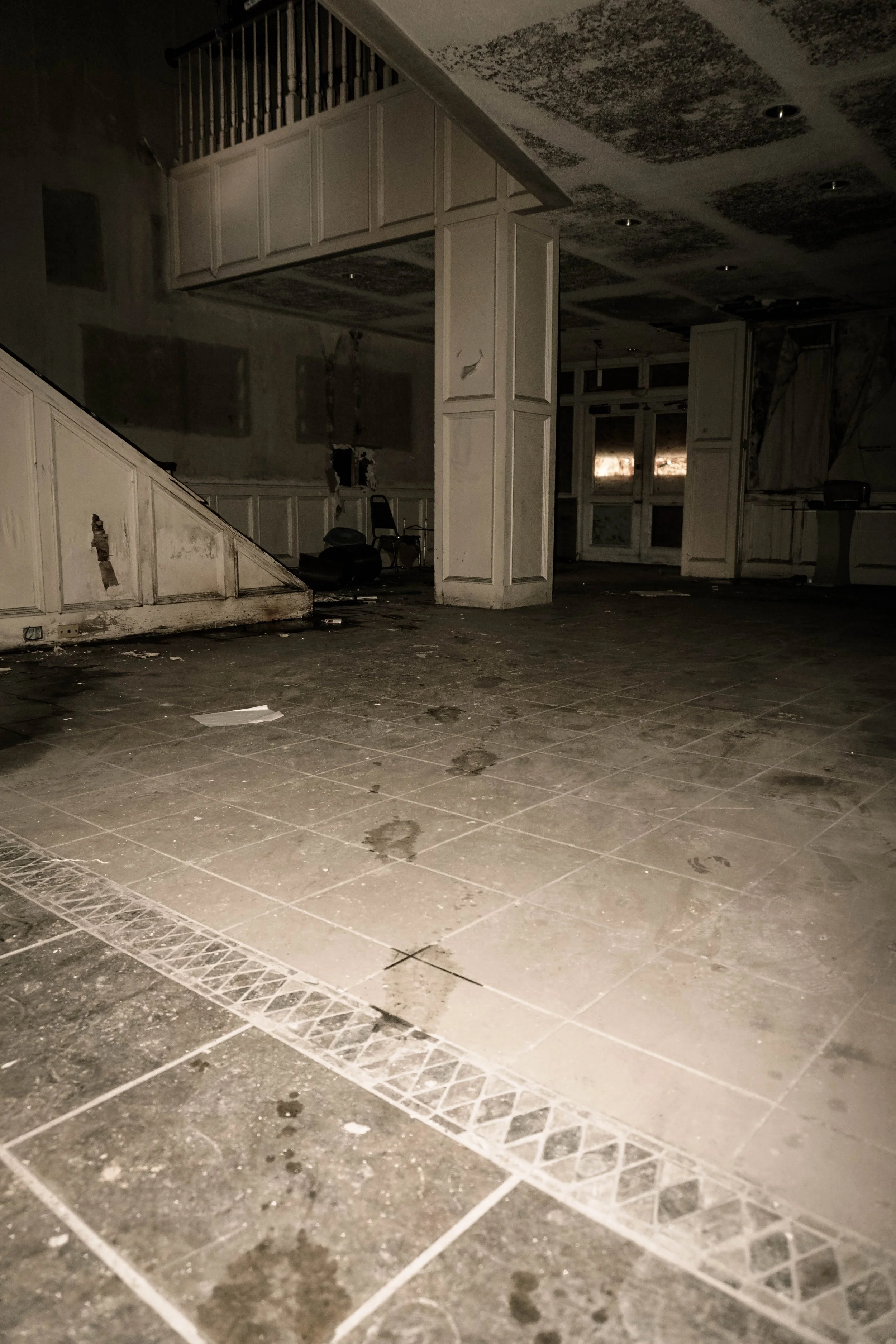 An abandoned, dilapidated interior space with dirty tiled flooring, peeling walls, and broken fixtures. The room appears empty and neglected.