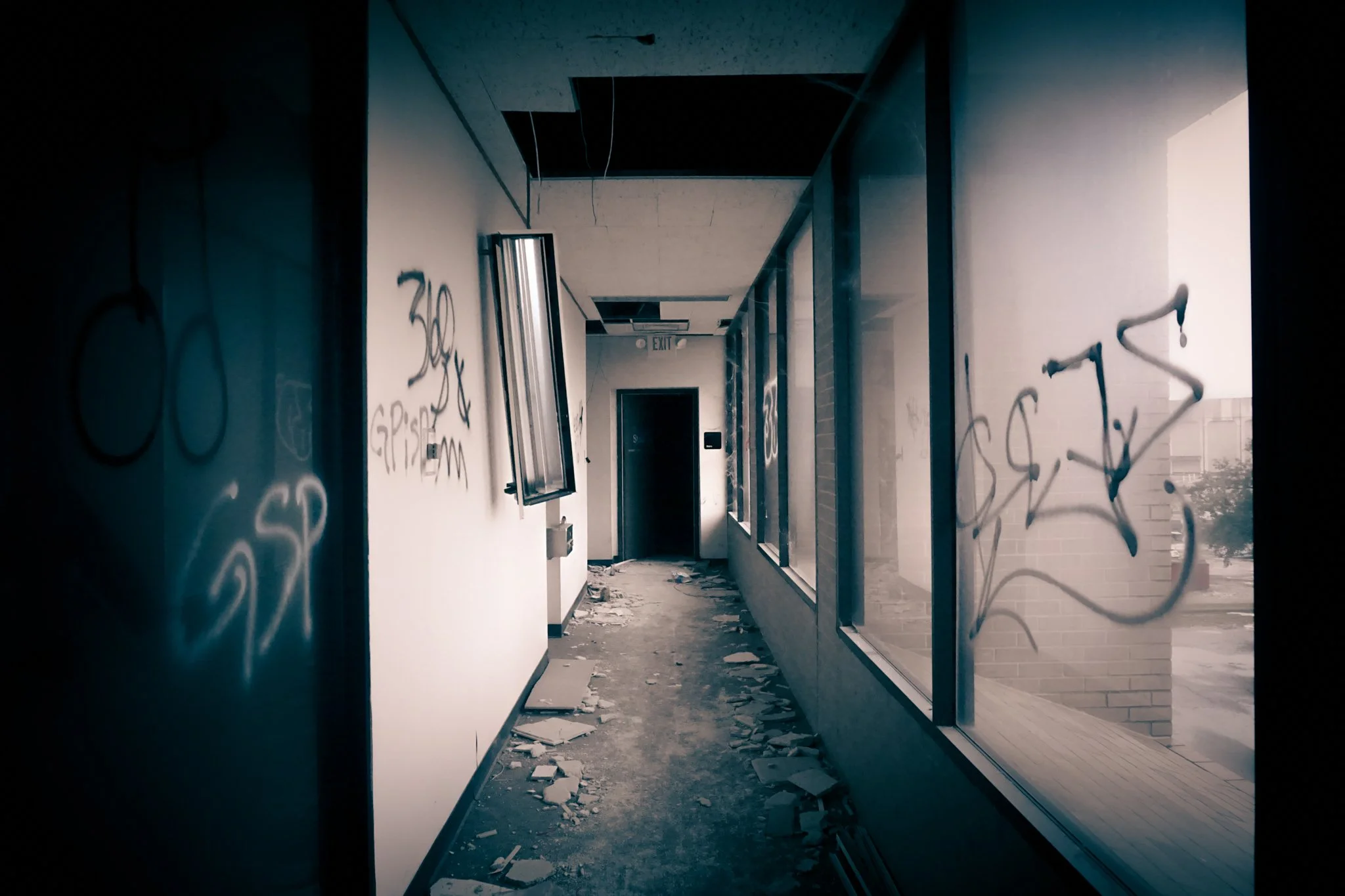 Abandoned hallway with debris on the floor, graffiti on the walls, broken window, and open ceiling panels, in a vacant building.