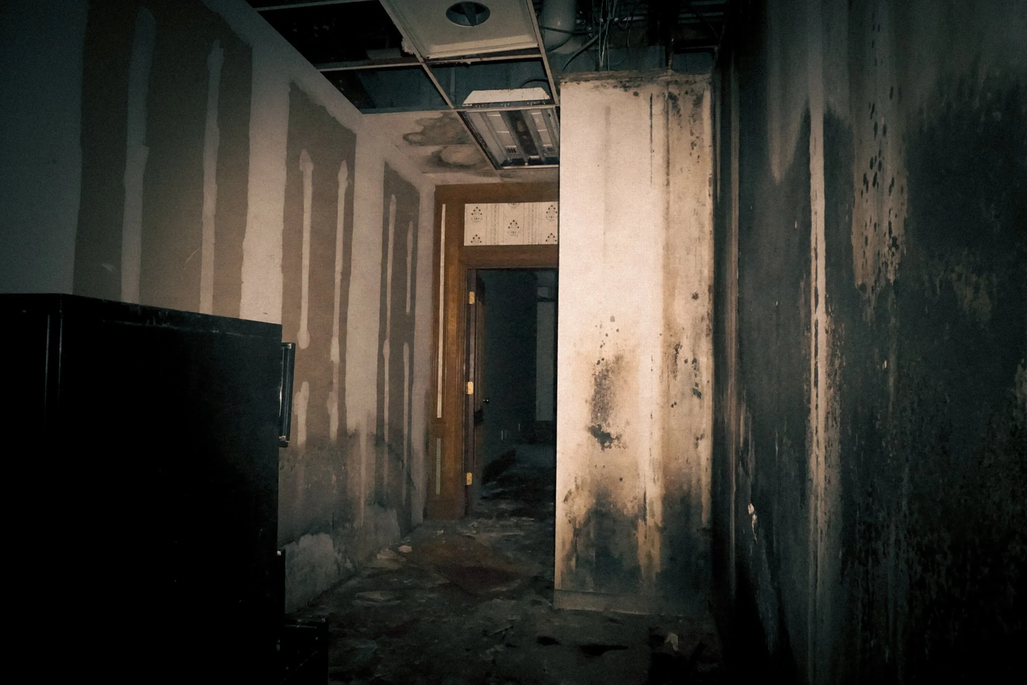 Interior of a building with burned and damaged walls, exposed ceiling framework, dark flooring, and a wooden door at the end of the hallway.