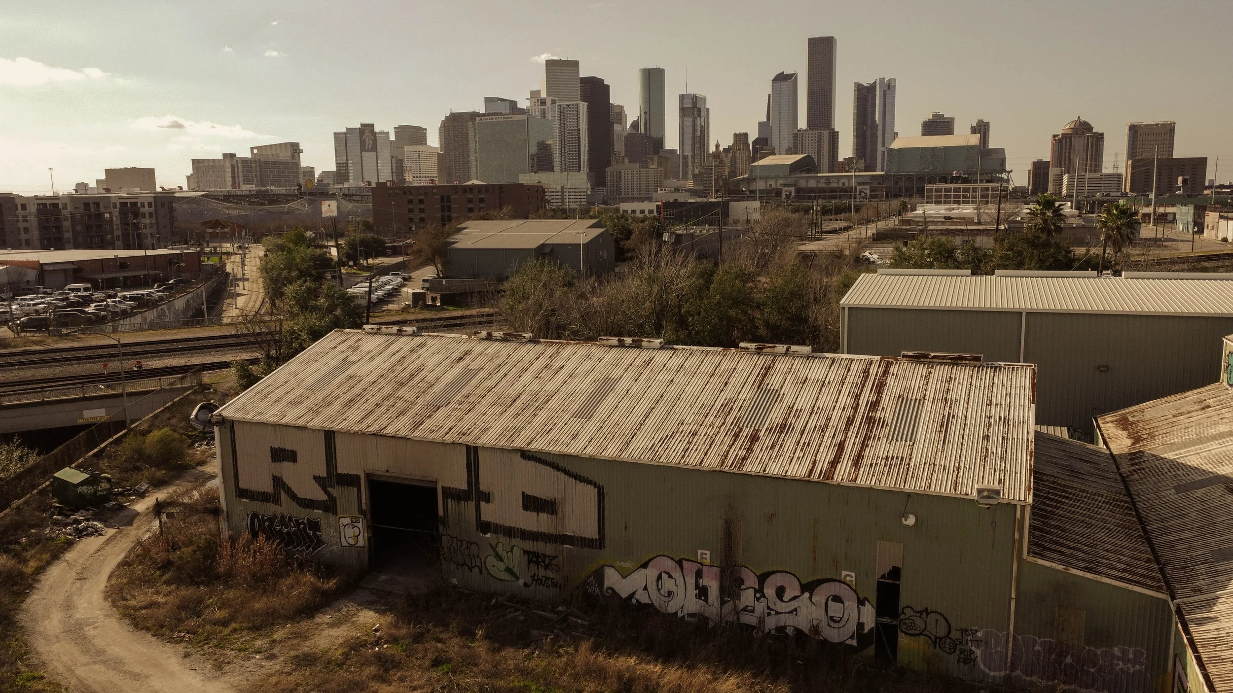 City skyline with tall skyscrapers in the background, an industrial warehouse with graffiti on the walls in the foreground, and a dirt path alongside parked cars and trees.