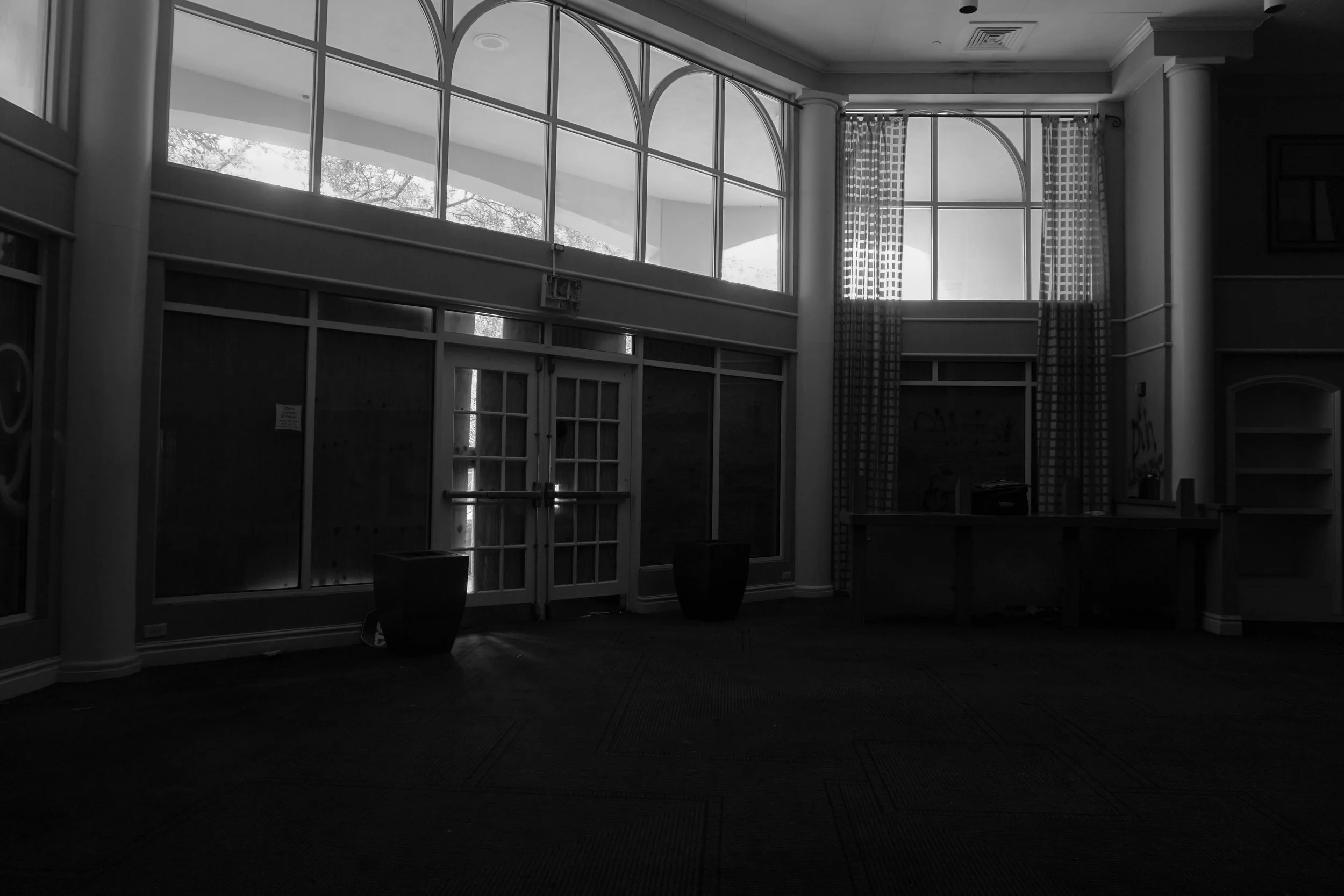 Empty interior space with large arched windows, curtains, glass doors, and a reception desk, illuminated by natural light.