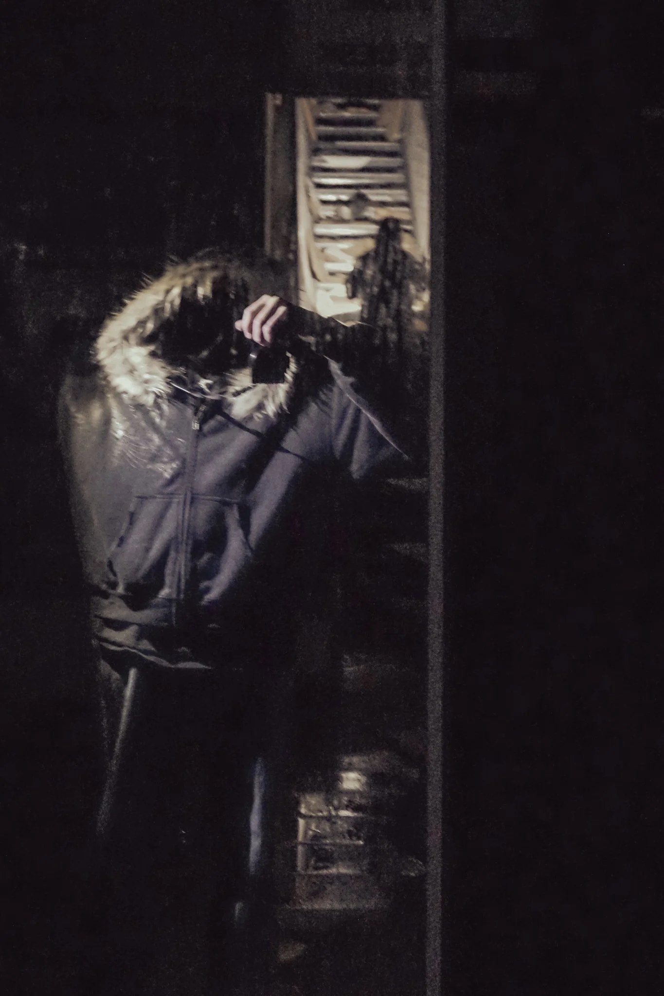 Person taking a mirror selfie in a dark room, wearing a black coat with a fur-lined hood, standing in front of a mirror with a wooden shelf or cabinet behind.