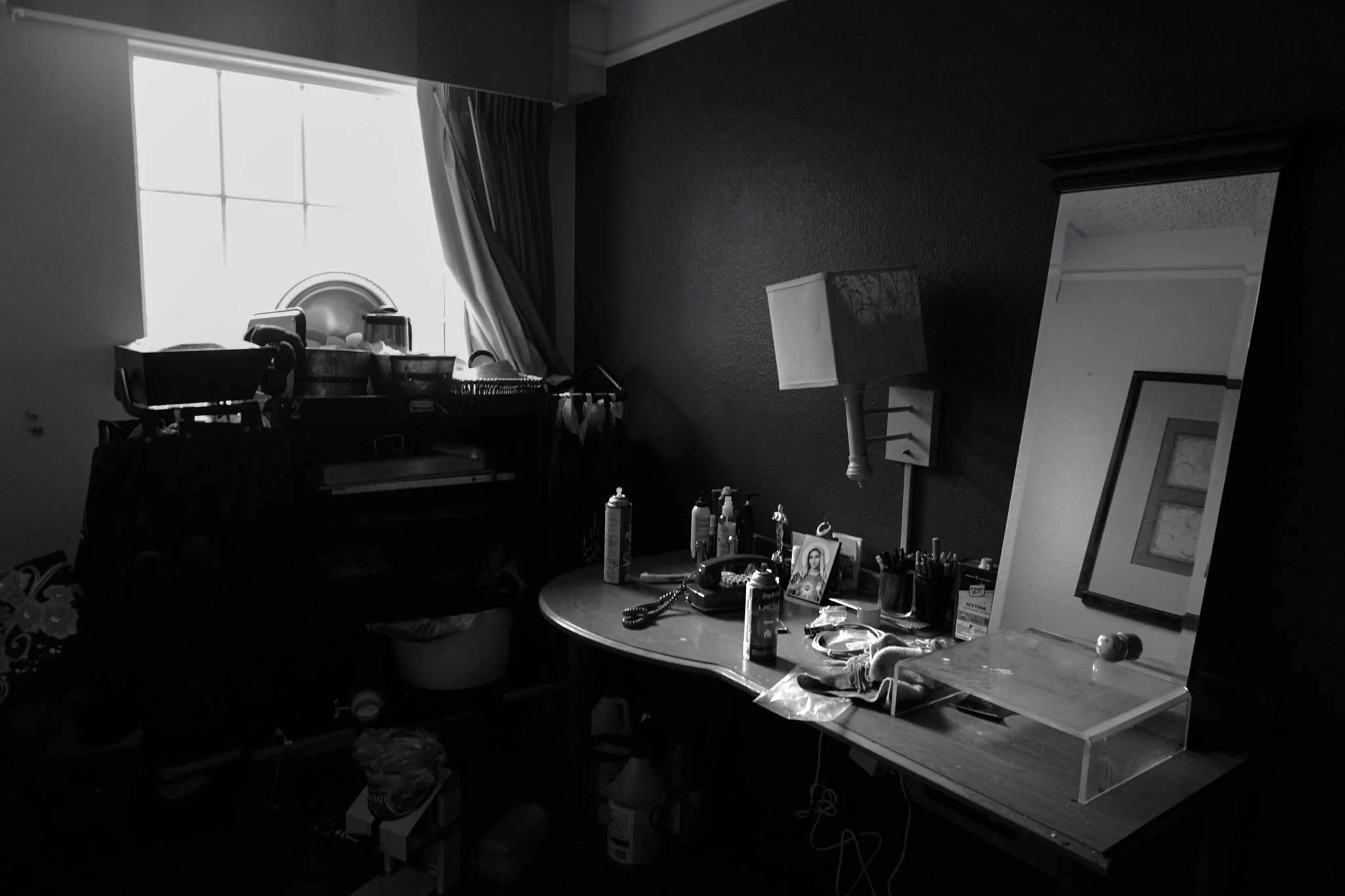 A cluttered desk beside a window with curtains, filled with various objects including bottles, framed pictures, and office supplies, and a mirror reflecting part of the room.
