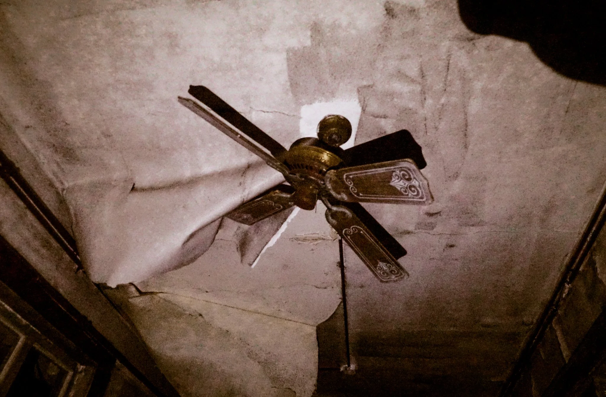 A ceiling fan with four blades hangs from a cracked and damaged ceiling with peeling paint.