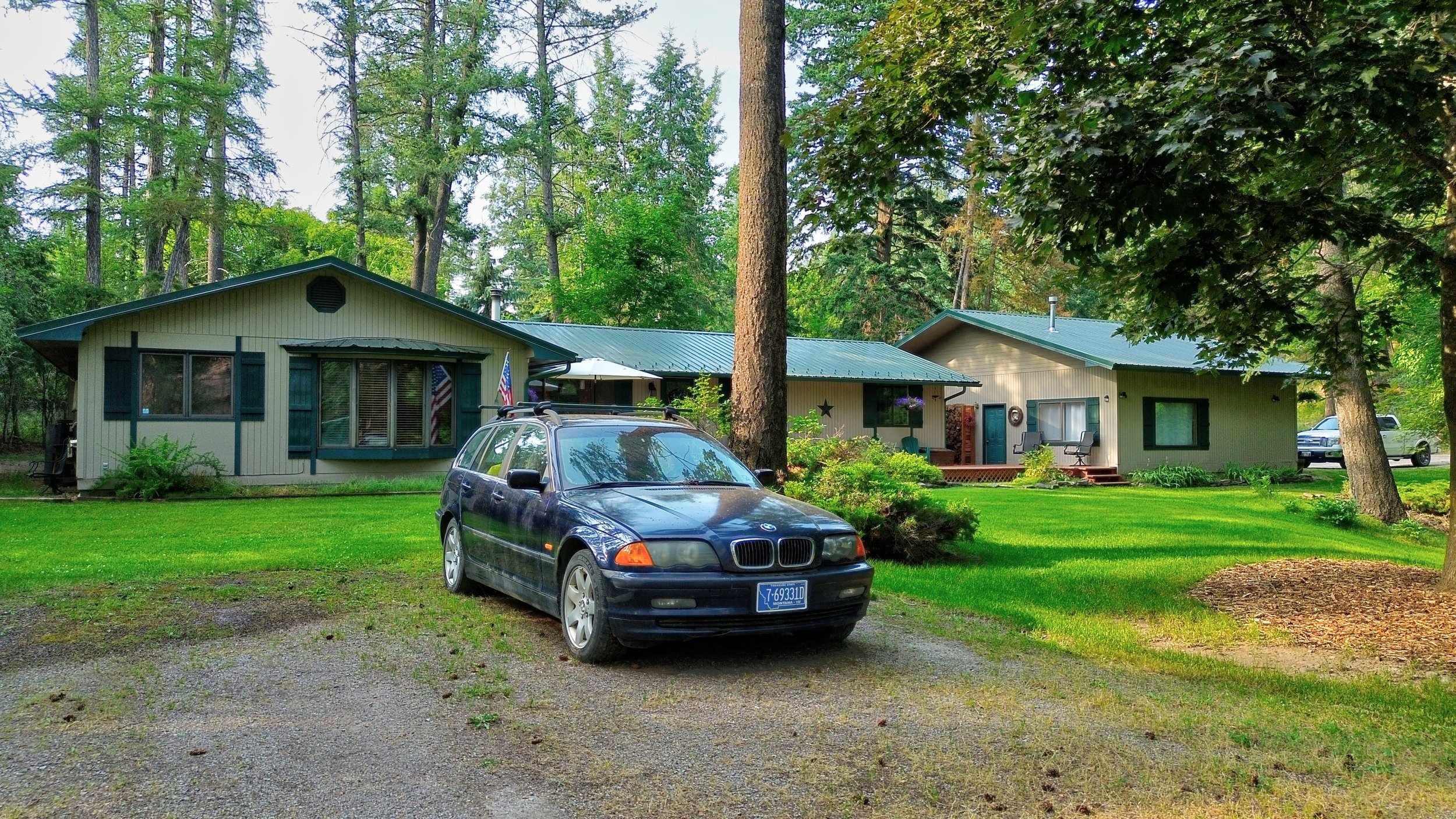 A house surrounded by tall trees and a well-maintained lawn, with two parked cars in the foreground and outdoor seating on the porch.