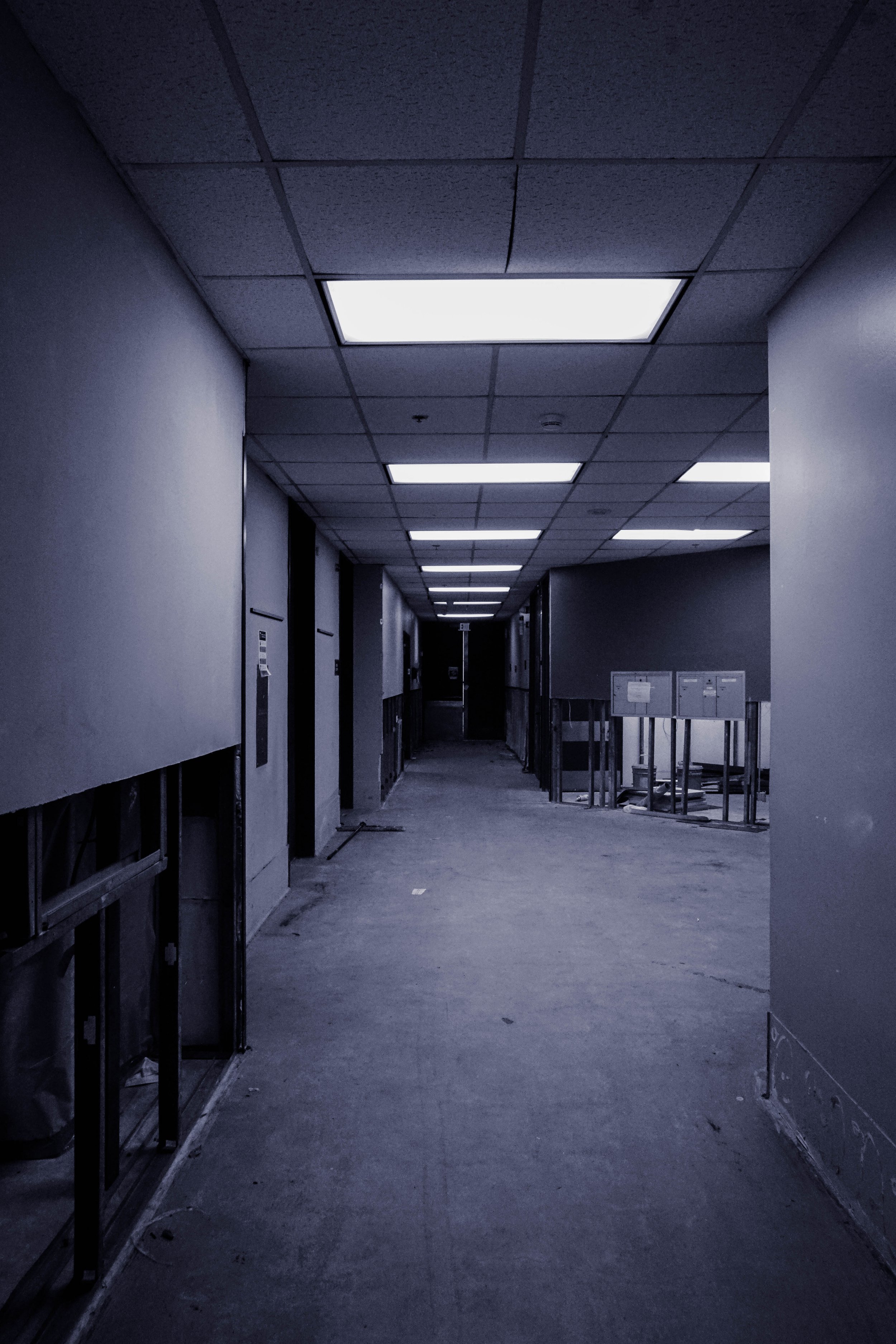A dimly lit empty corridor under construction with unfinished walls and ceiling panels, and bright ceiling lights.