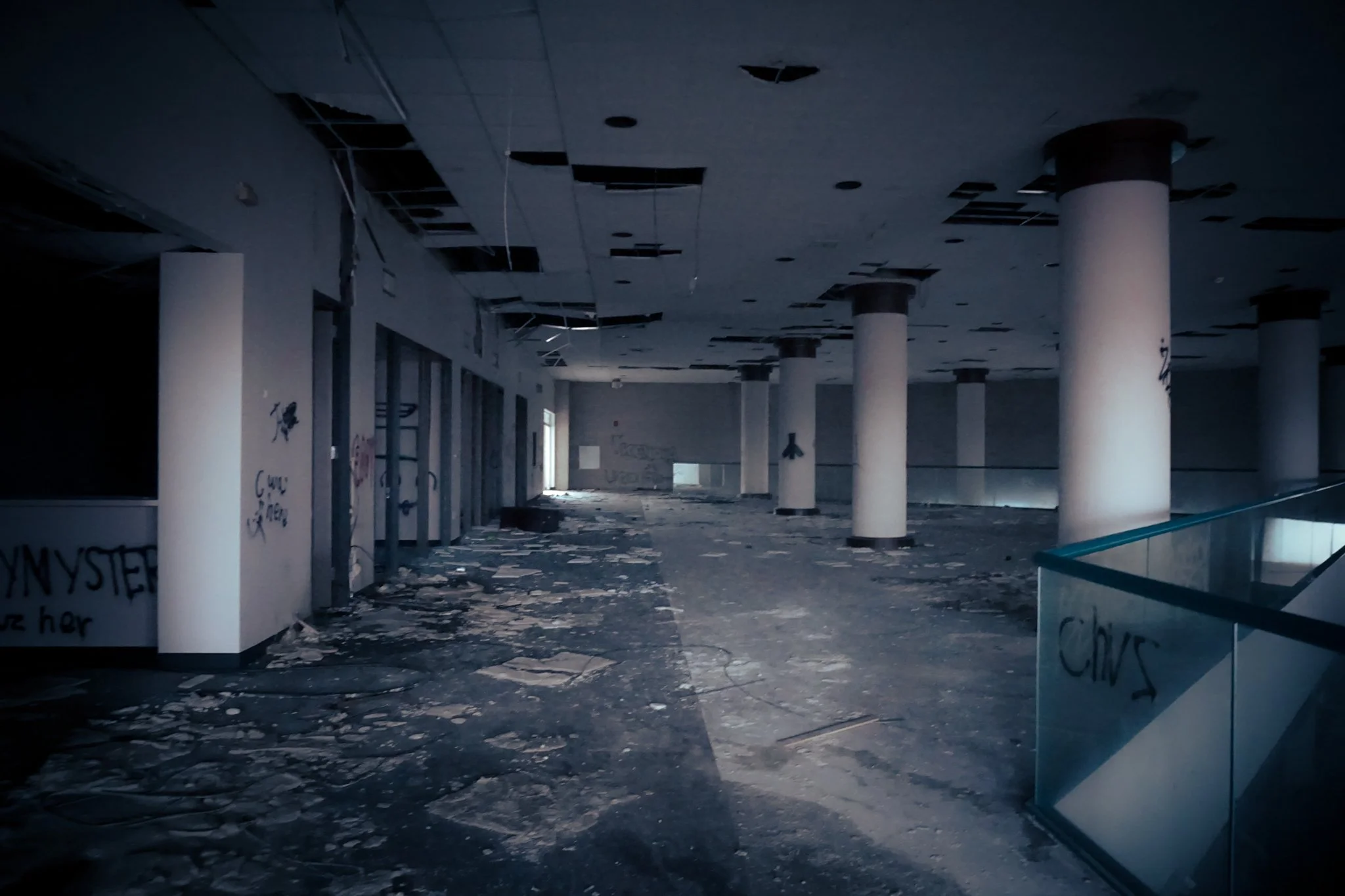 Interior of an abandoned building with a damaged ceiling, graffiti on the walls, debris on the floor, and broken glass railings.