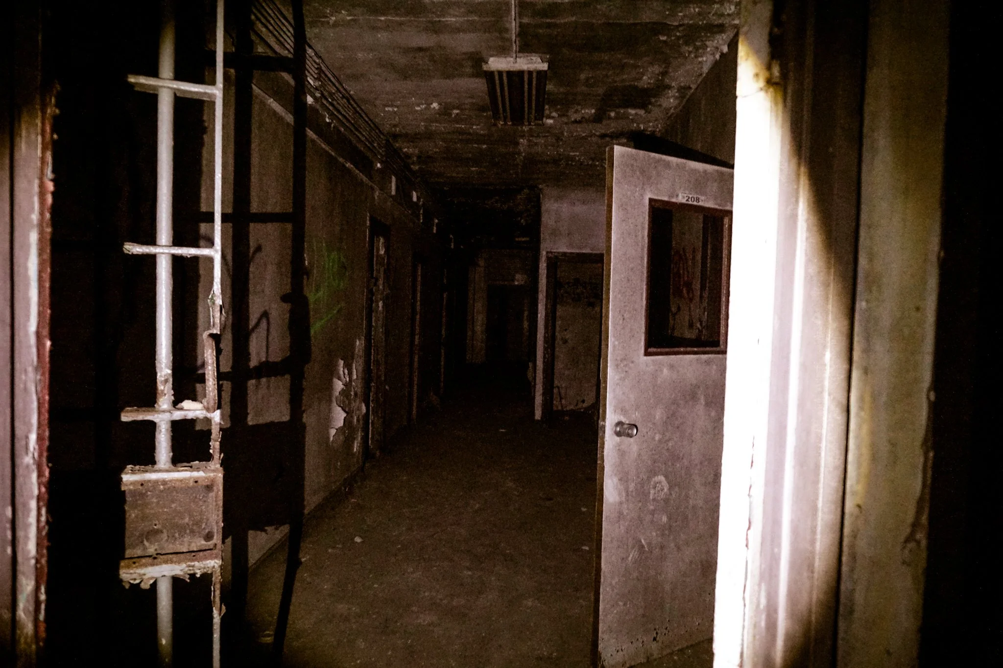 Dark, abandoned hallway with open door on right and industrial fixtures on ceiling.