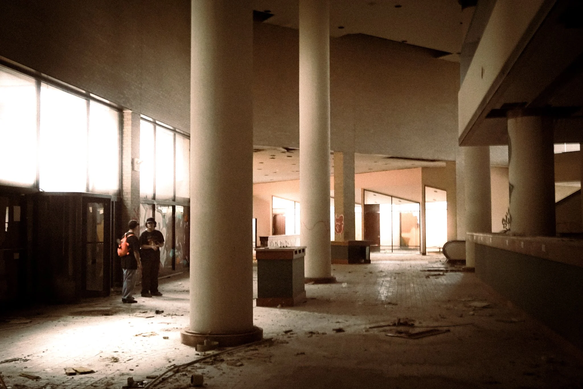 An abandoned, dimly lit interior of a building with large vertical pillars and broken windows. Two people are standing together on the left. The floor is dirty with debris scattered around.
