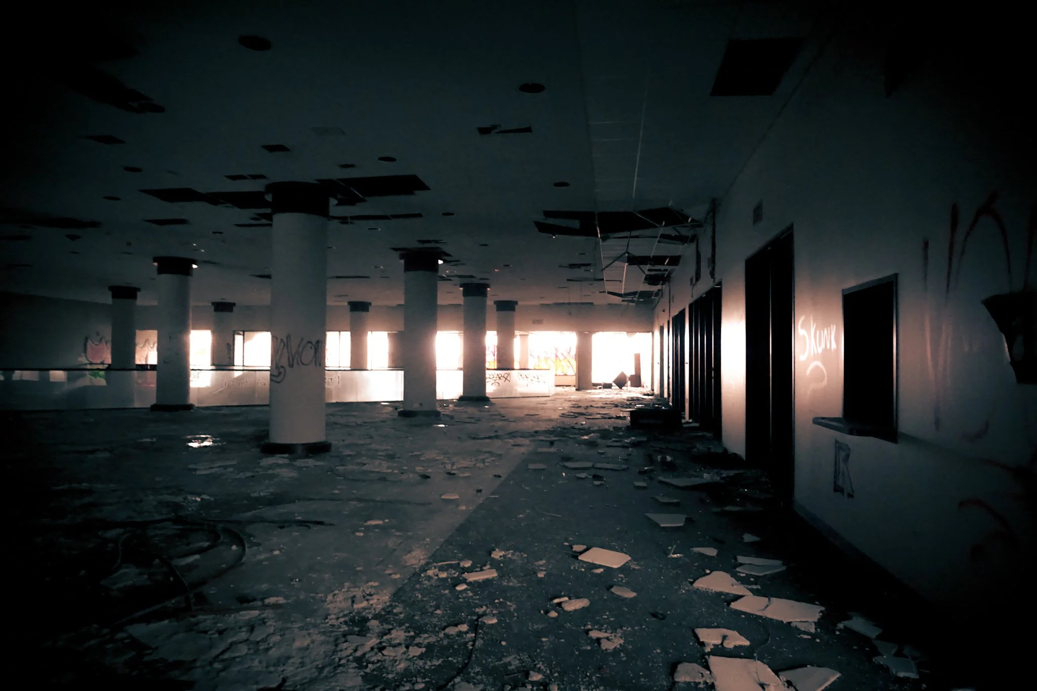 Dark, abandoned interior with broken ceiling panels, debris on the floor, and sunlight coming through the windows in the back.