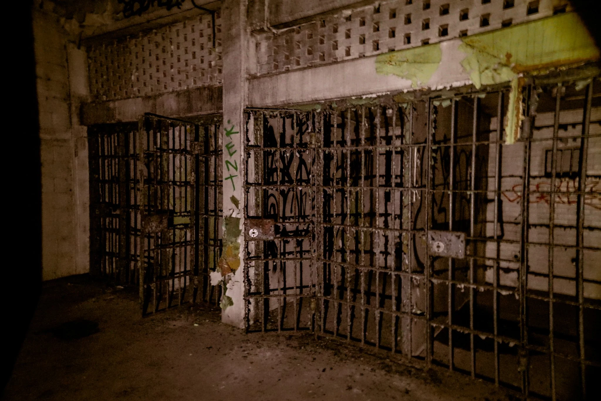 An old, rusty gate with vertical bars and a latch in a dilapidated, graffiti-covered wall, possibly a jail cell or abandoned building.