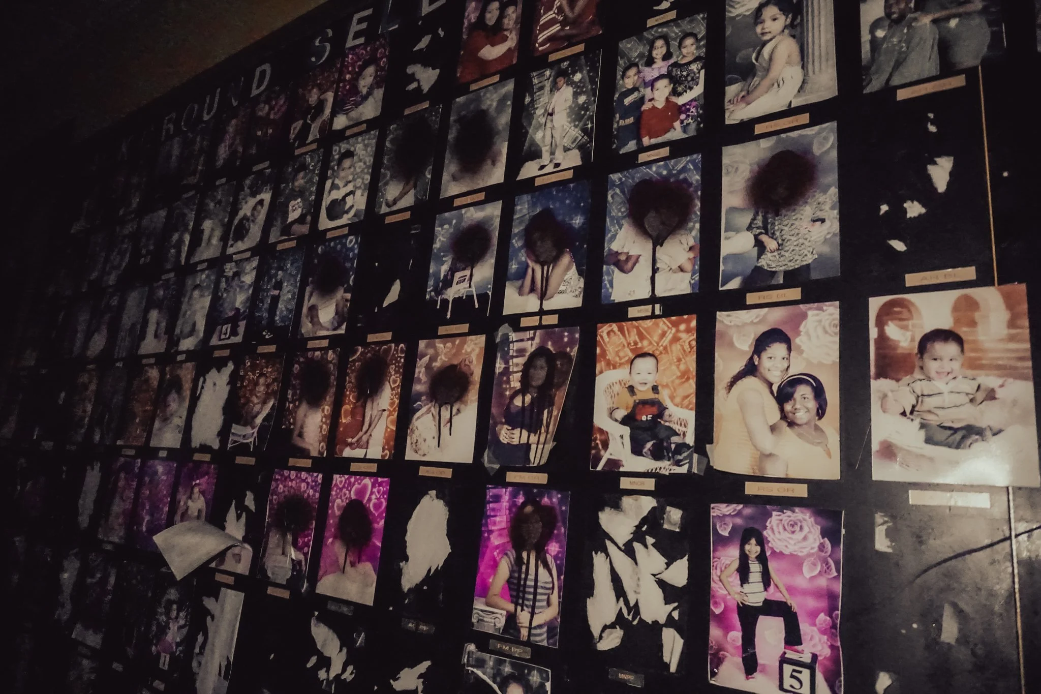 A wall display of multiple photographs of children, some in groups and some solo, arranged in rows with labels under each picture. The photos include a variety of poses and backgrounds, with some children smiling and others with neutral expressions.