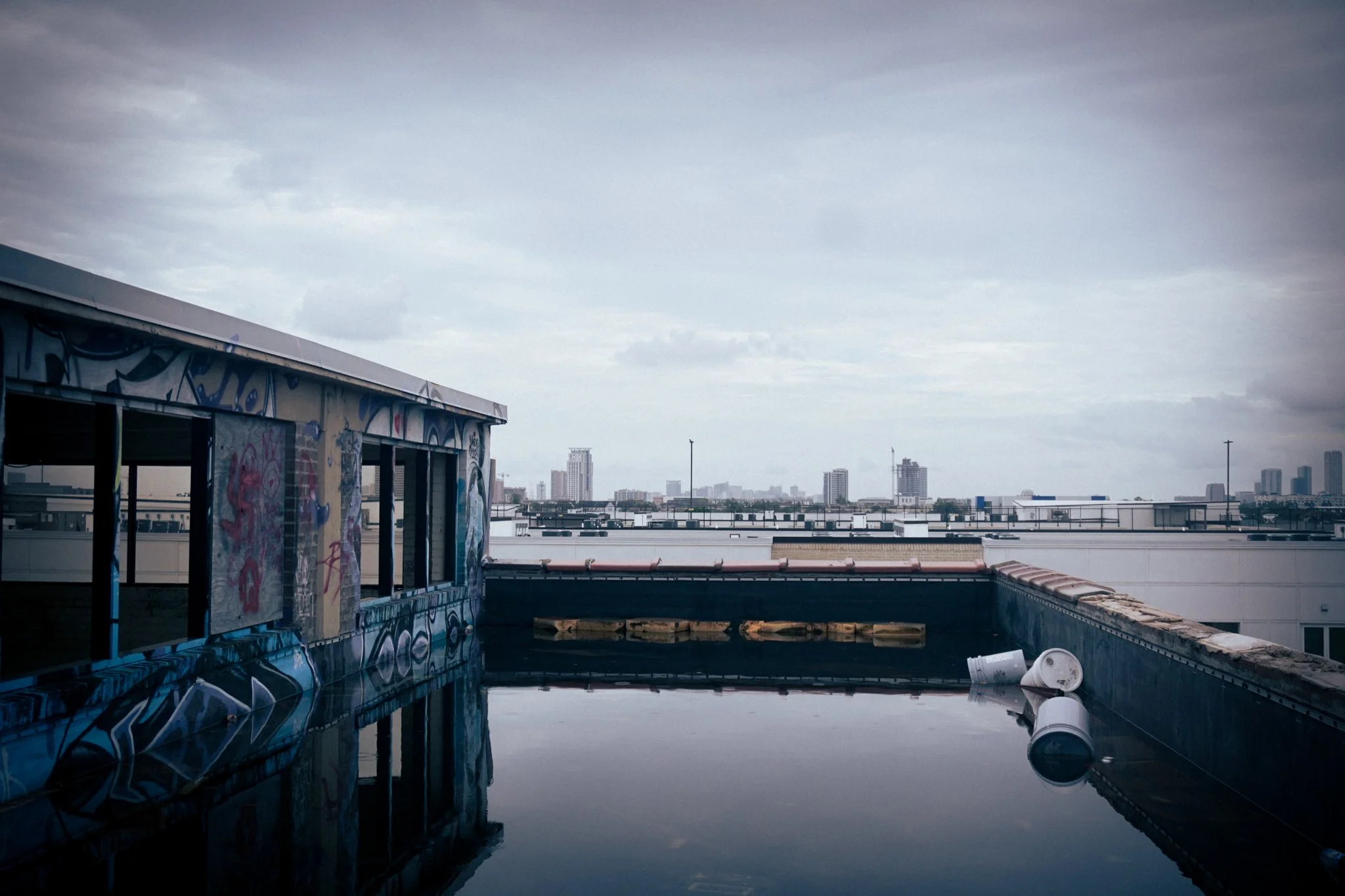 Rooftop with Graffiti walls and a pool of water, city skyline in the background, cloudy sky.