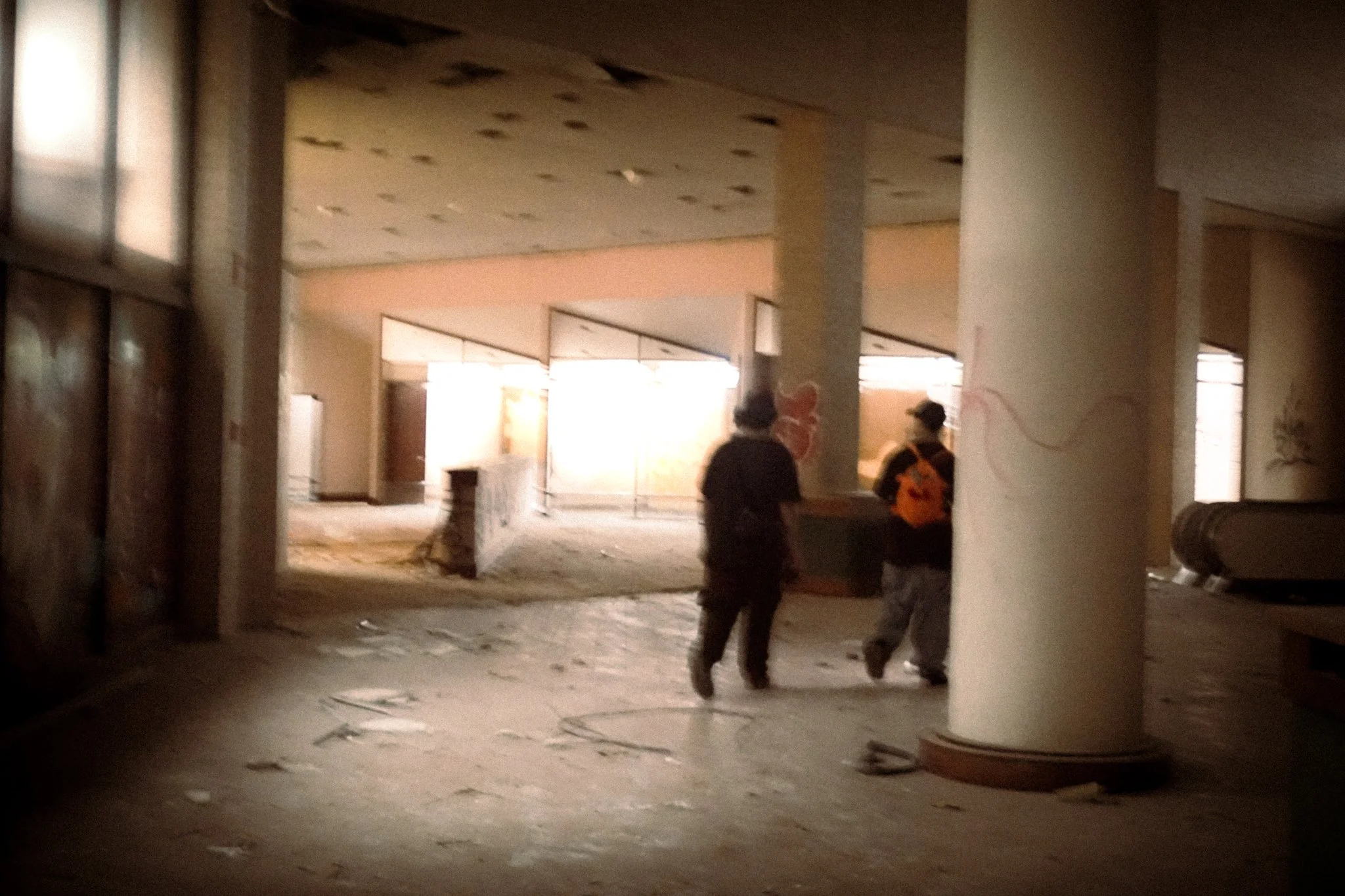 Two people walking through an abandoned, dilapidated building with debris on the floor and sunlight streaming in from the open area ahead.