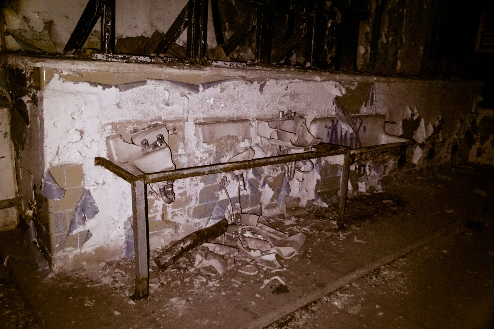 Three old soap sinks mounted on a rusted metal frame, behind a destroyed brick and plaster wall in a dilapidated building.