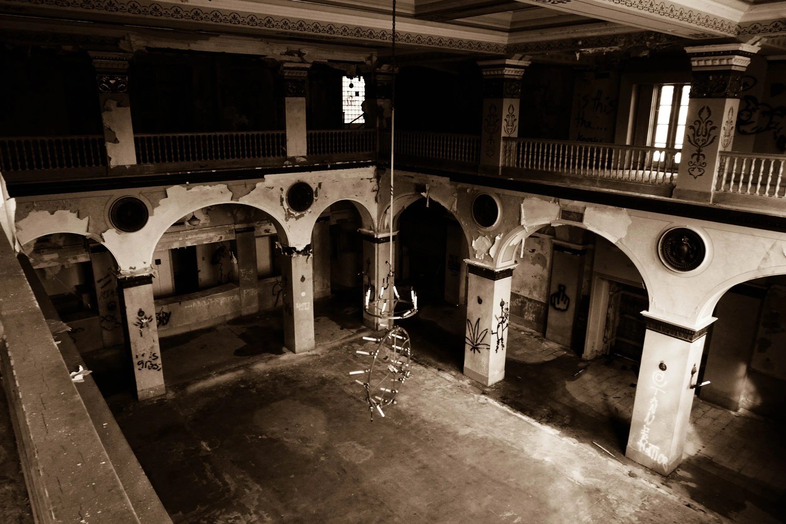An abandoned, graffiti-covered interior of a dilapidated two-story building with arched doorways, a chandelier hanging from the ceiling, peeling walls, and broken windows.