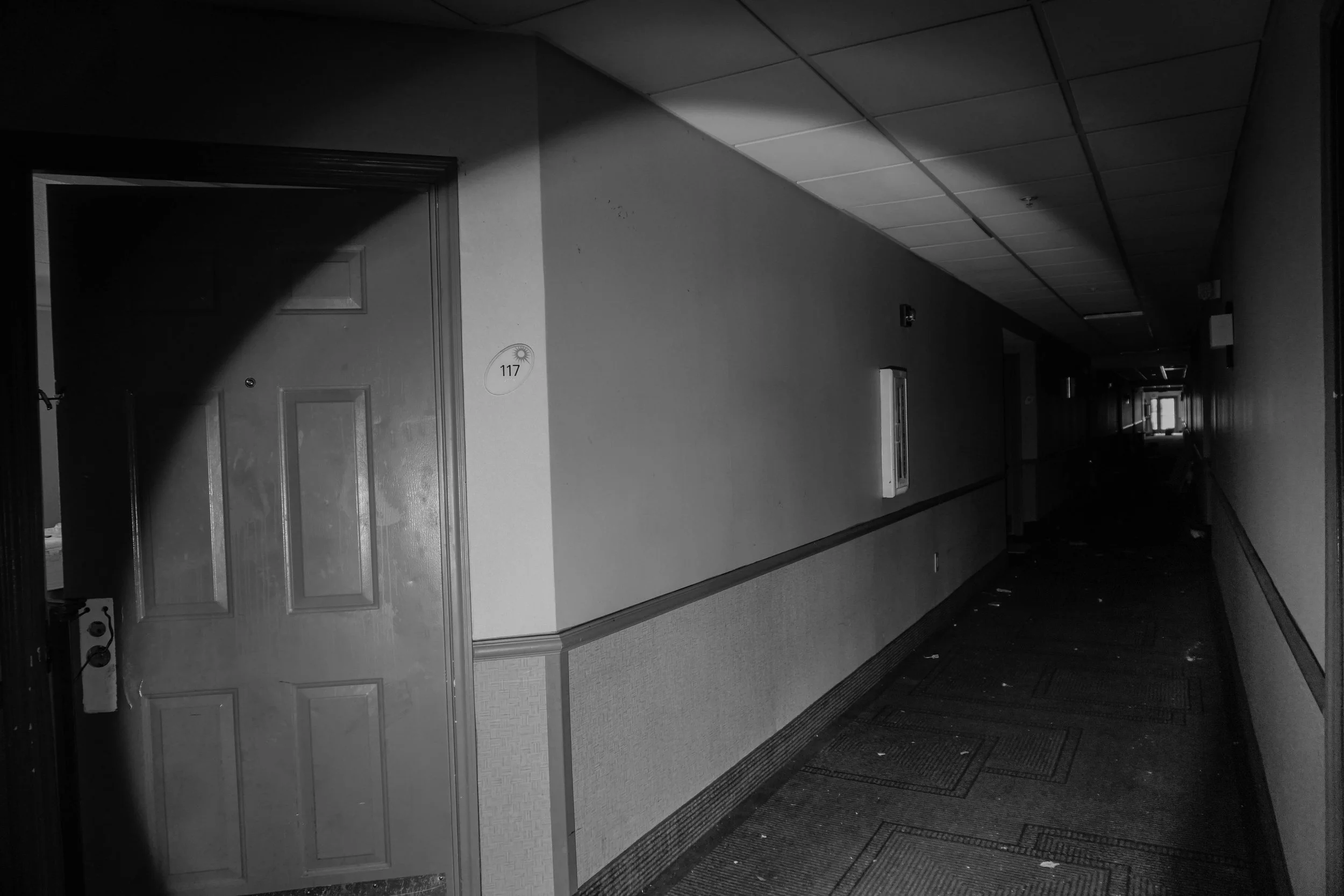 Dimly lit hallway with closed door labeled 117 on the left and a narrow corridor extending into the distance. Carpeted floor with debris scattered.