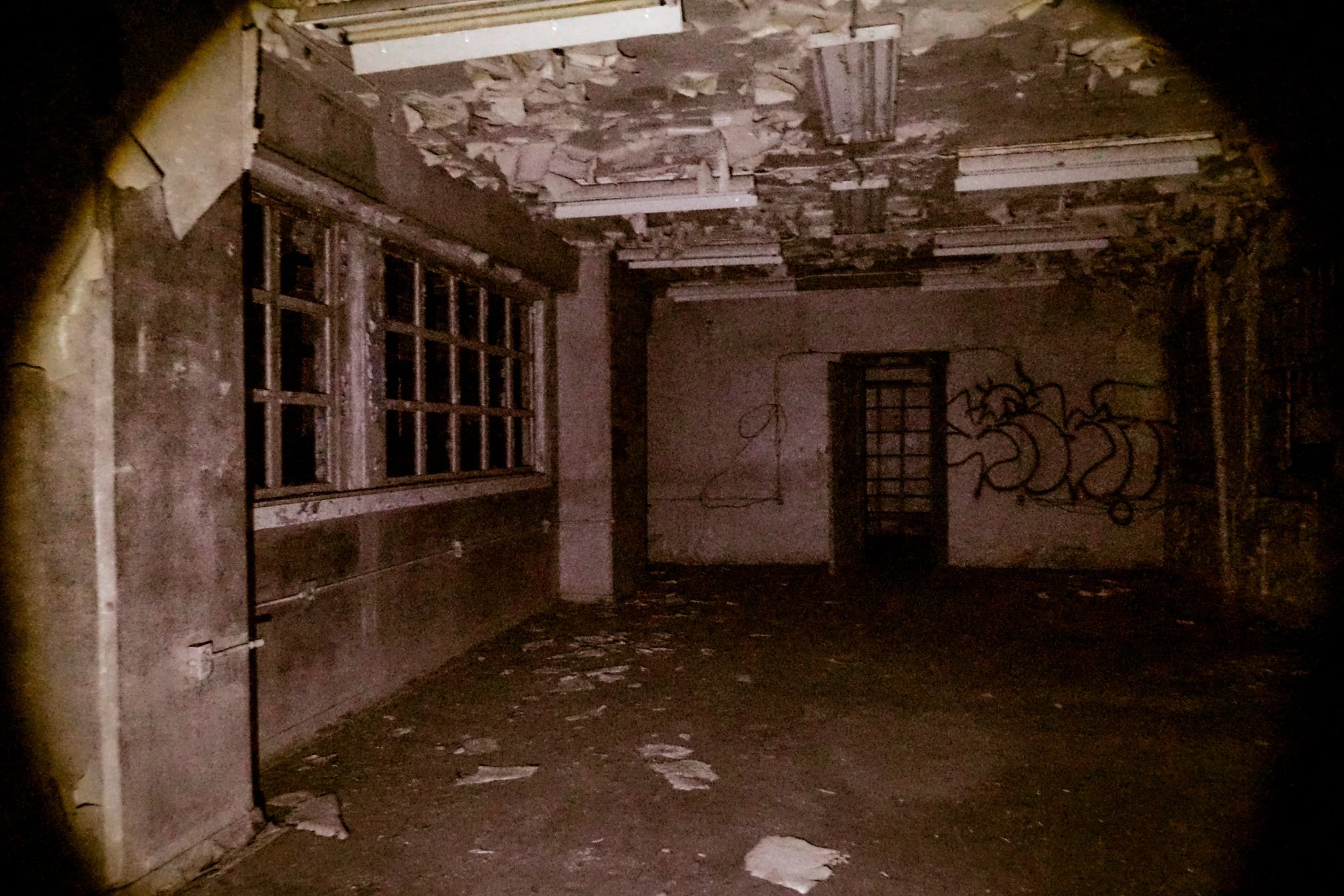 An abandoned and dilapidated room with peeling paint, broken ceiling tiles, graffiti on the walls, a large window with missing glass, and debris scattered on the floor.