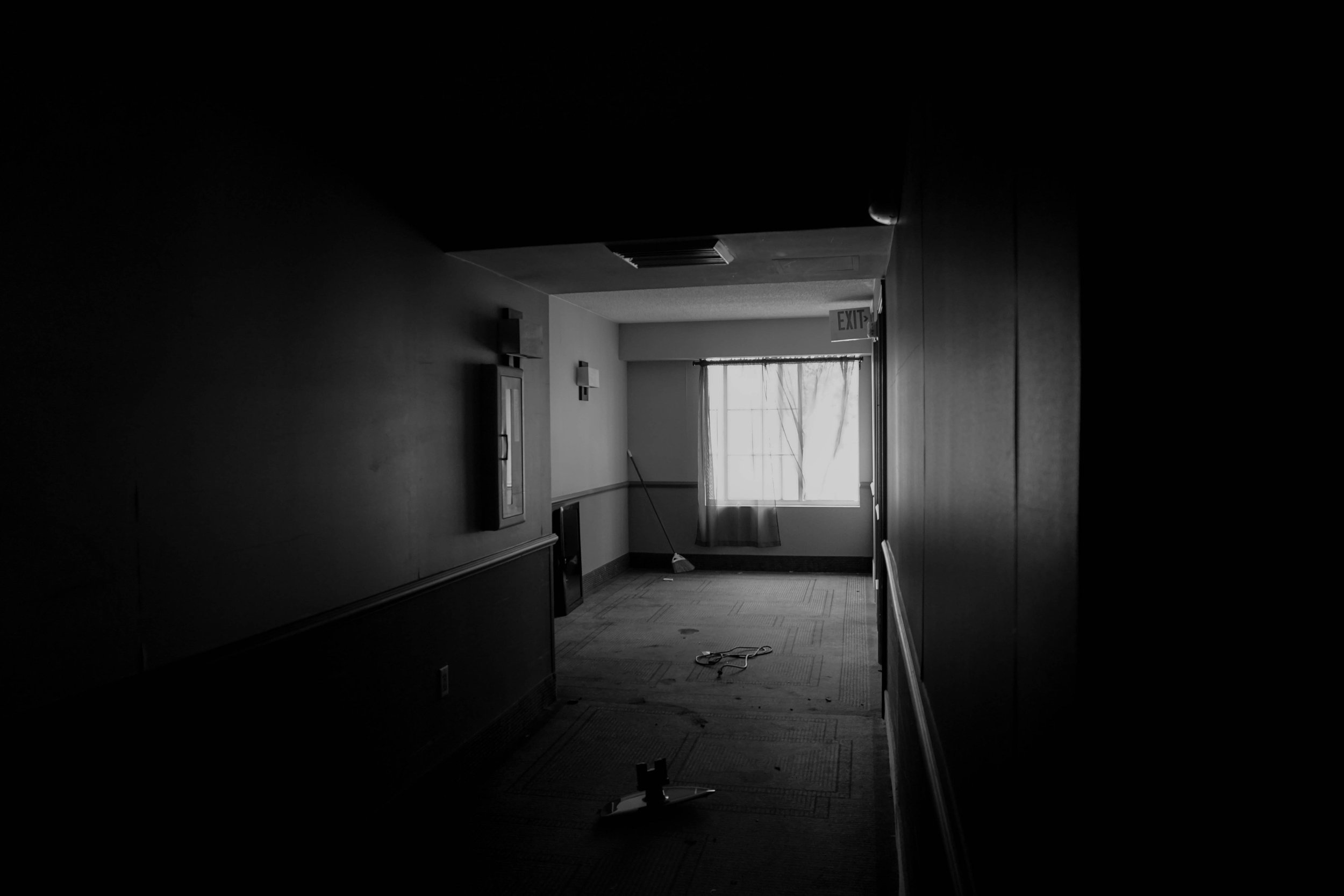 Empty hallway in a building with a window at the end, a mop leaning against the wall, and some scattered objects on the floor.