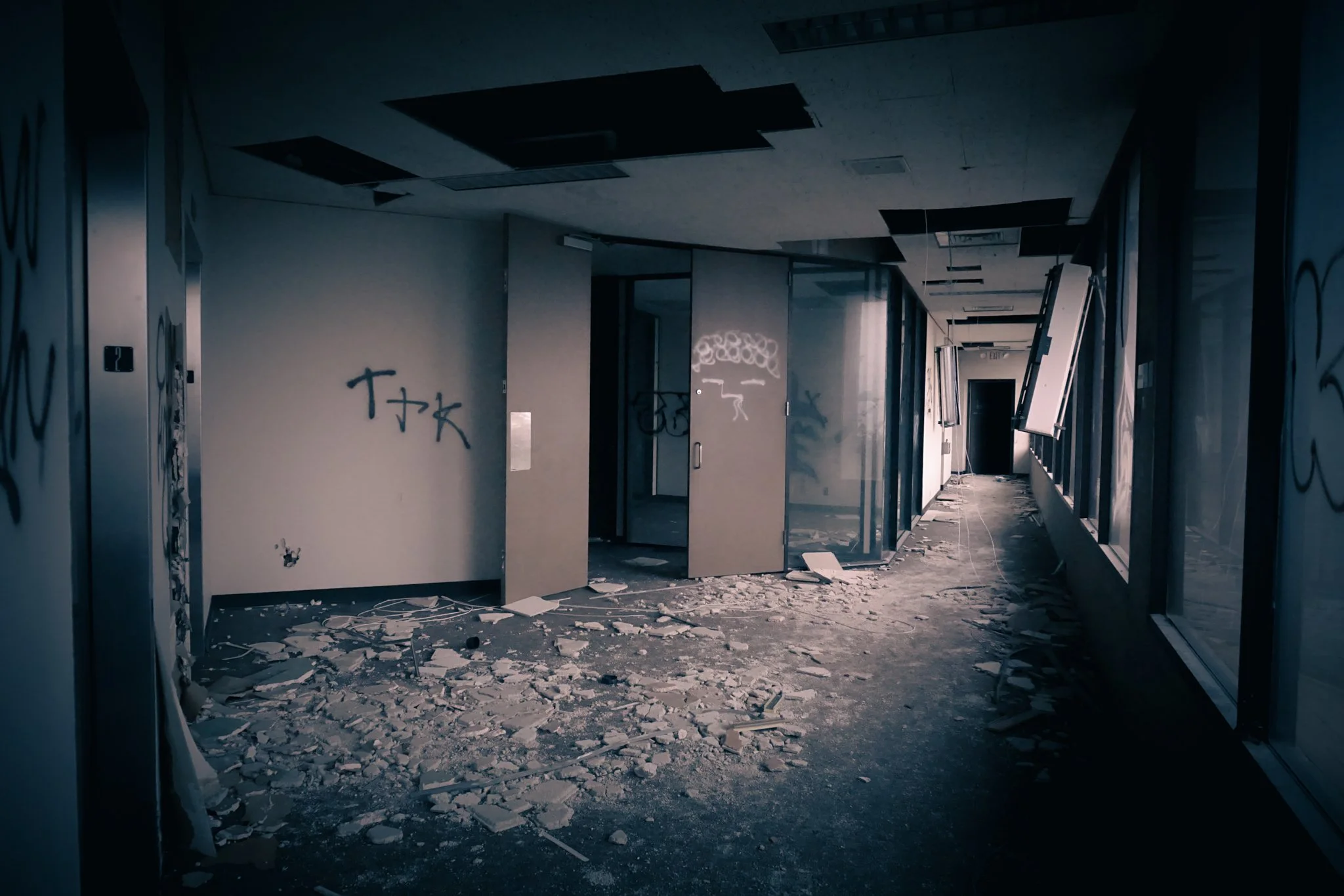 Abandoned interior space with broken ceiling tiles, graffiti on walls, and debris scattered across the floor.