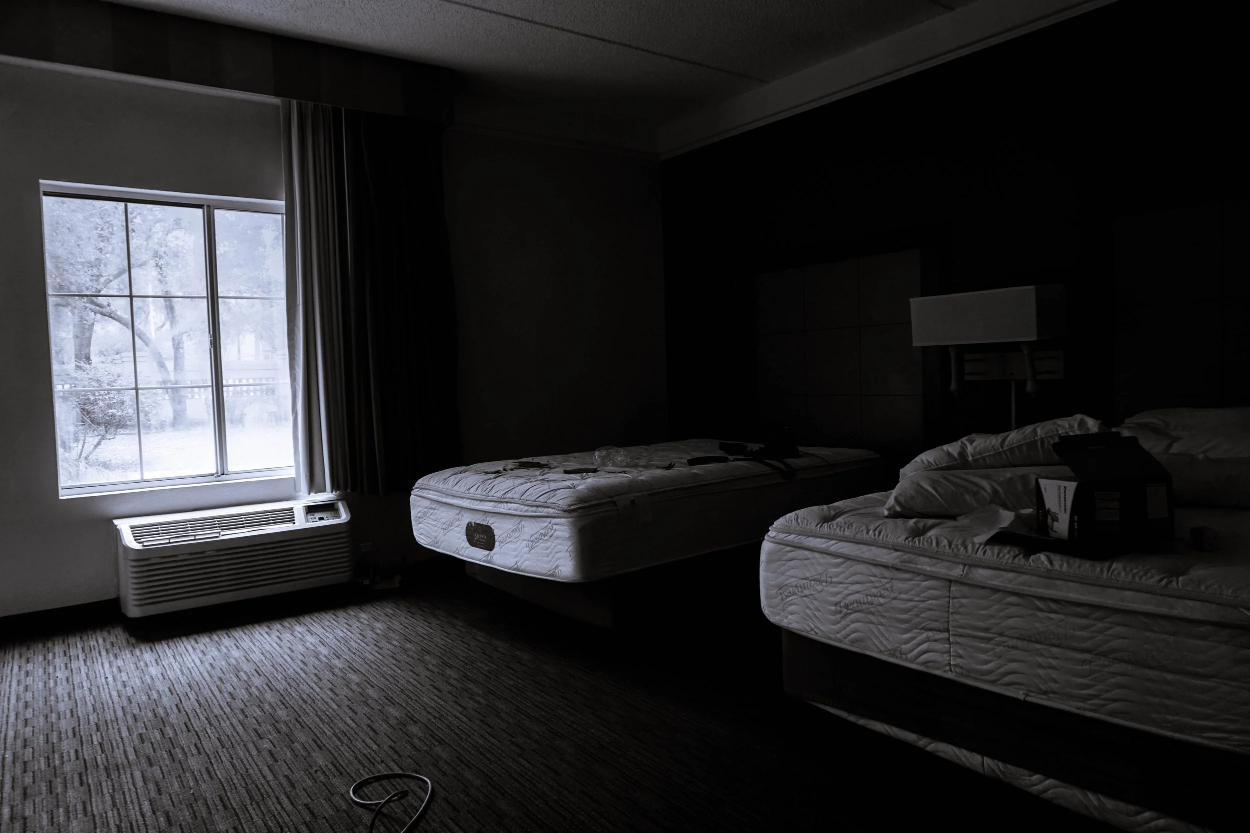 A dark hotel room with two unmade beds, a window showing a snowy landscape outside, and an air conditioning unit beneath the window.