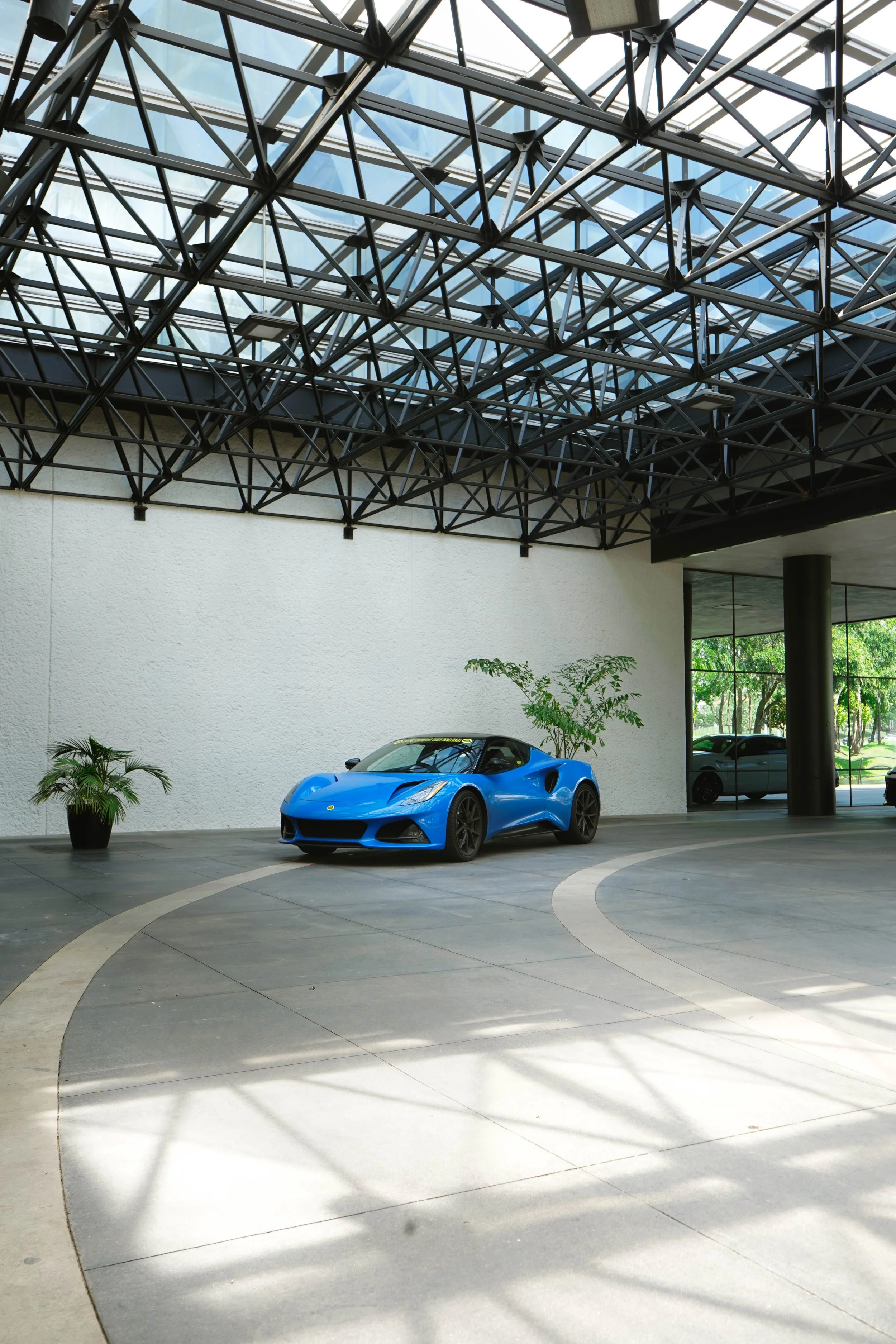 A bright blue sports car parked inside a modern building with a glass and steel ceiling structure. There are a few potted plants and a large window with natural light coming through.