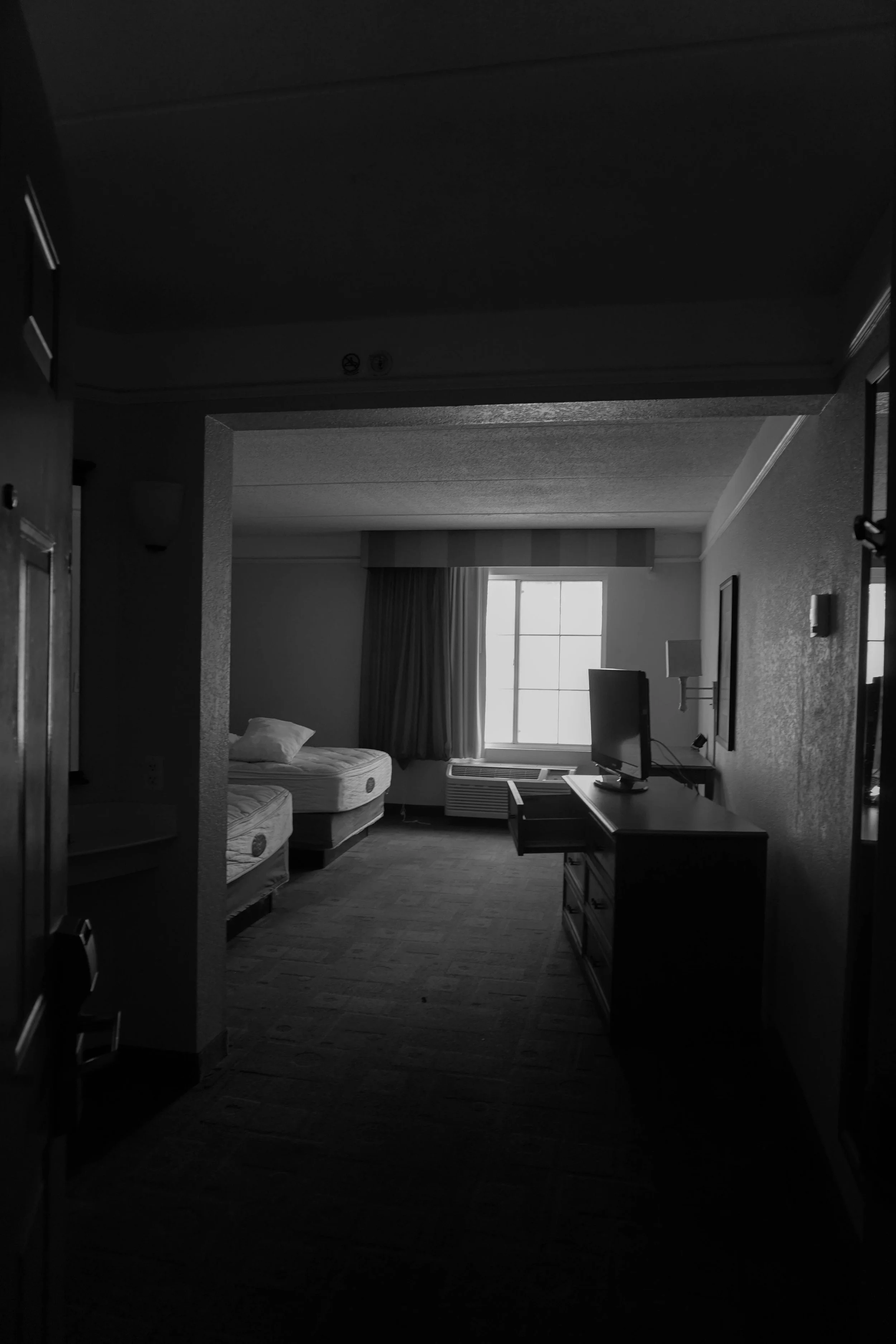 A black and white photo of a hotel room with two beds, a window with curtains, a TV on a dresser, and an air conditioning unit below the window.